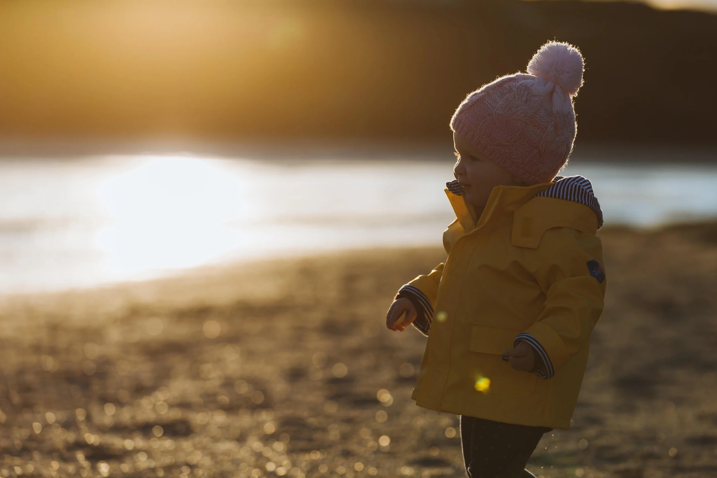 Child wearing a pink knit hat and yellow jacket standing on a beach during sunset, facing to the right.