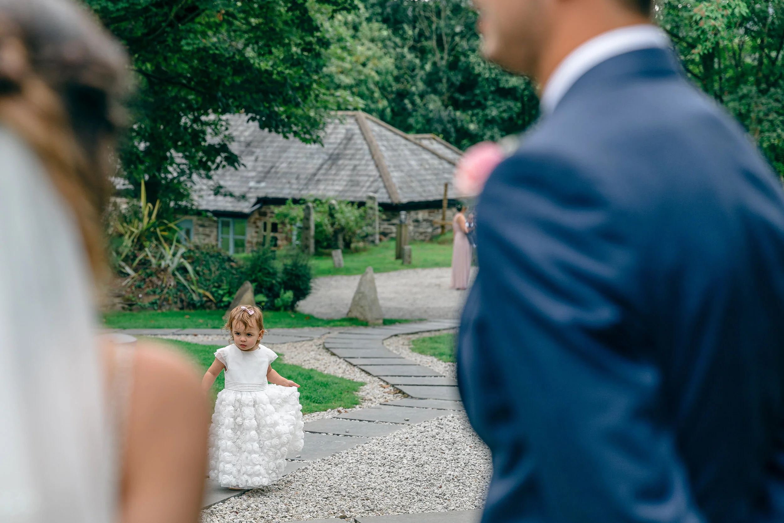 A young girl in a white dress walking on a gravel and stone pathway outdoors, with a woman in a wedding dress partially visible in foreground, and a man in a suit also partially visible. In the background, there is a rustic stone house and lush green