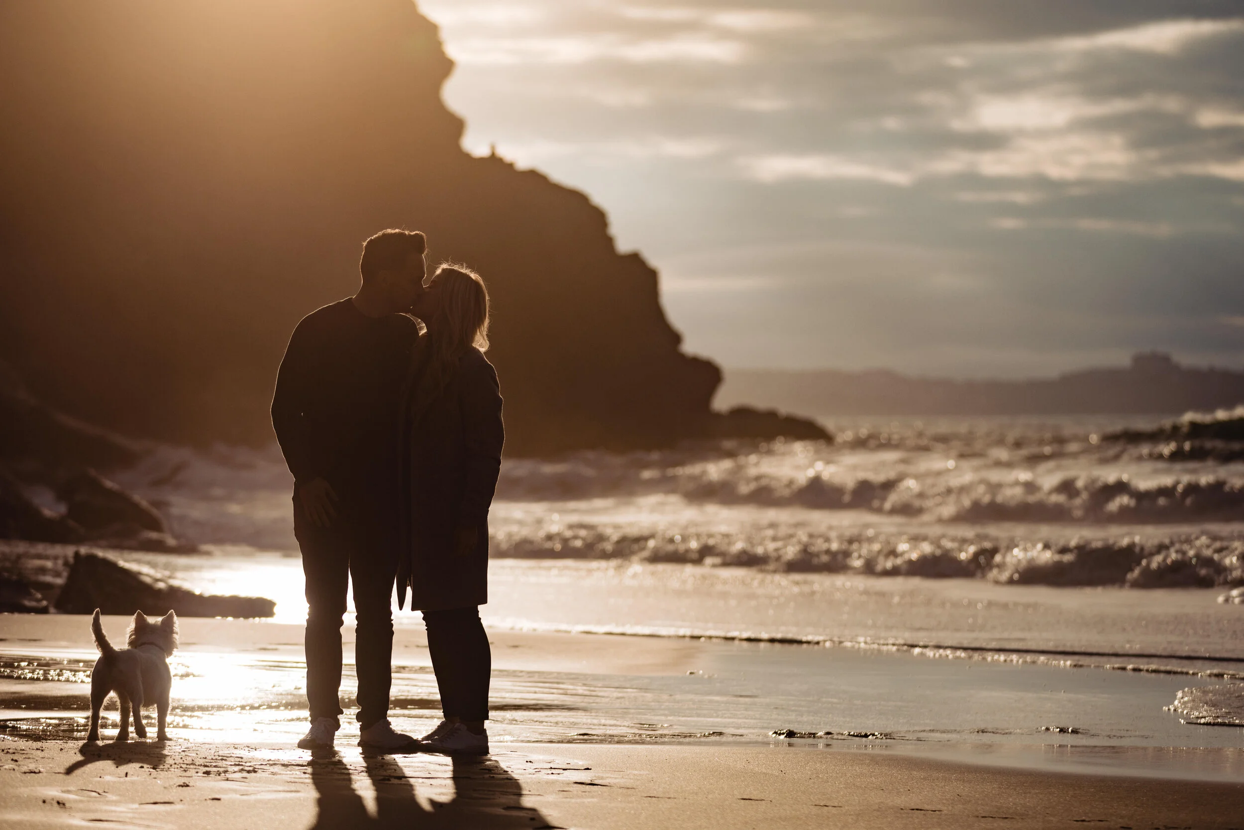 A couple kissing on the beach with a dog nearby during sunset, with cliffs and ocean in the background.