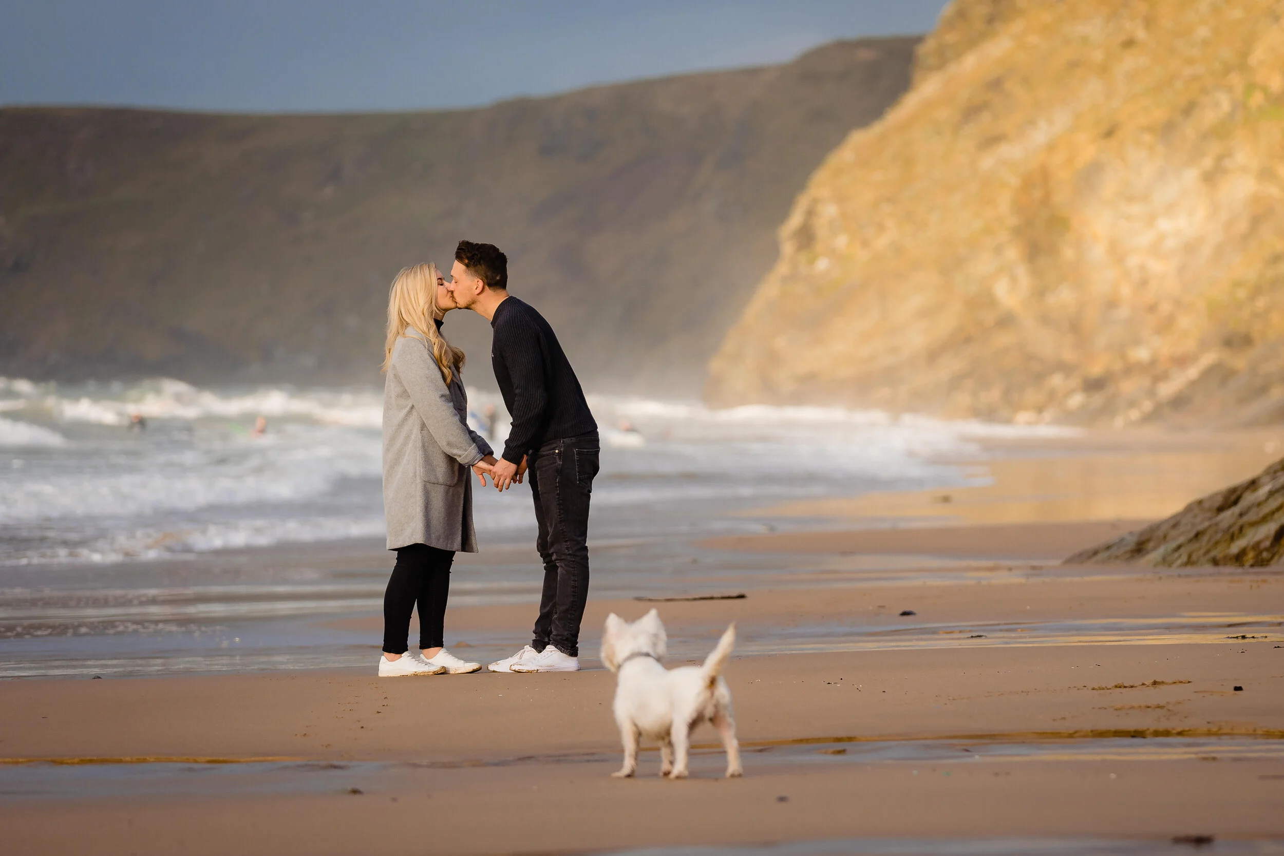 A couple sharing a kiss and holding hands on a beach with a dog in the foreground, cliffs in the background.