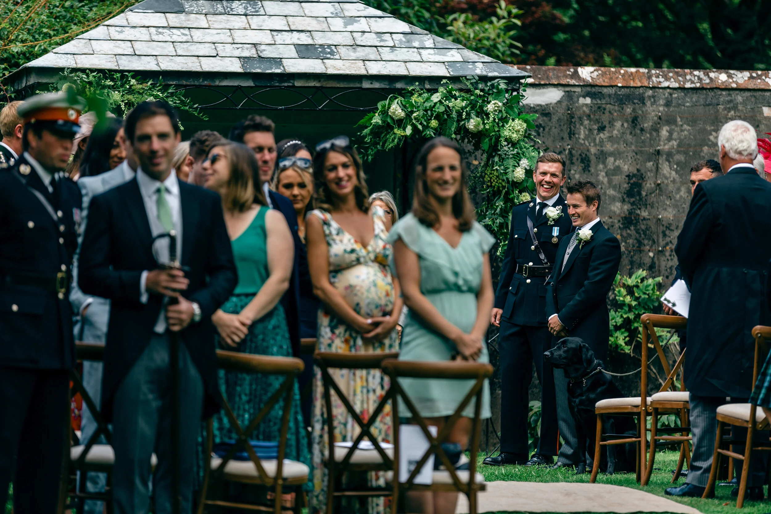 People attending an outdoor wedding ceremony, standing in front of a floral arch. Some are dressed in military uniform, others in formal attire, with a mix of men and women.