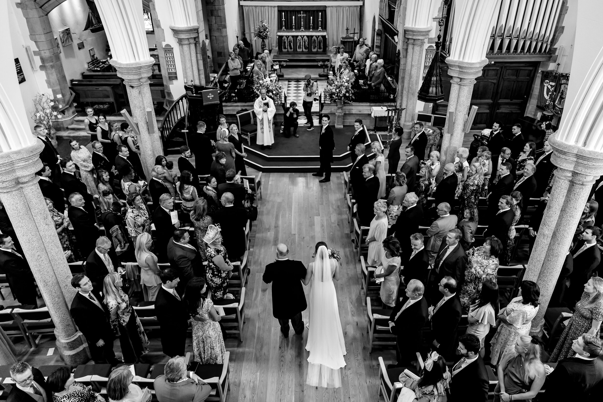 Black and white photo of a wedding ceremony inside a church. The bride, wearing a long white dress and veil, is walking down the aisle with a man. Guests are seated on either side, watching the couple. The church has tall columns, rows of pews, and a