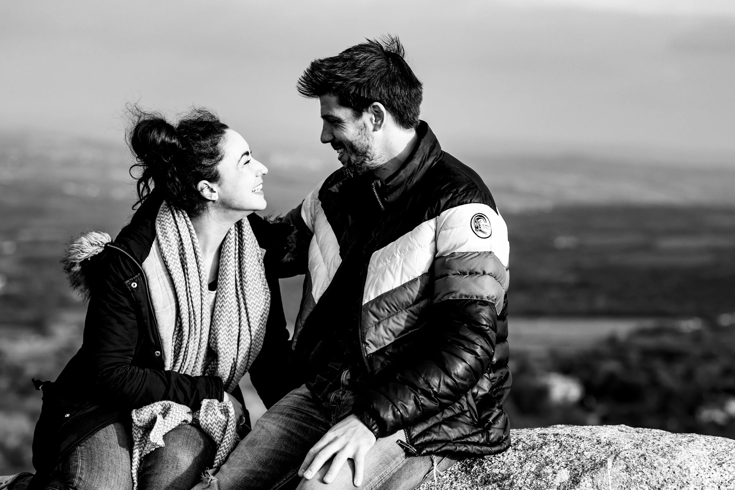 A young woman and a man sitting outdoors on a rock, smiling and looking at each other, with a blurred landscape in the background.