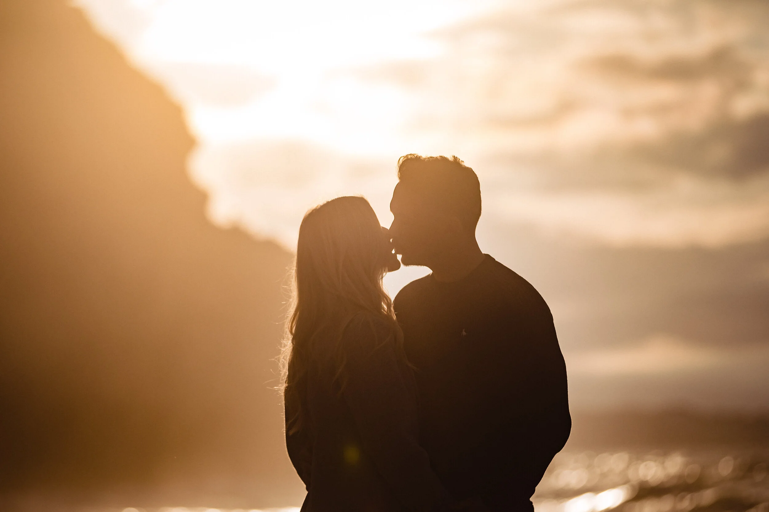 Silhouette of a couple kissing on the beach at sunset with the ocean and sky in the background.