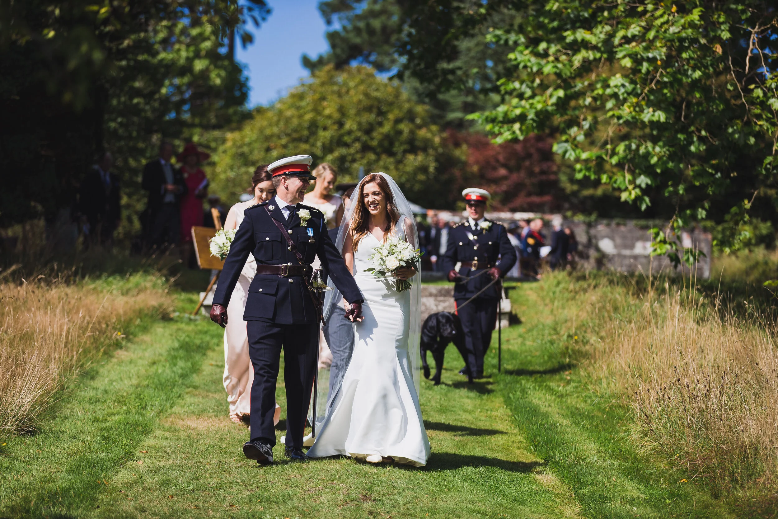 A wedding procession on a grassy path with wedding guests and armed police officers, surrounded by trees on a sunny day.