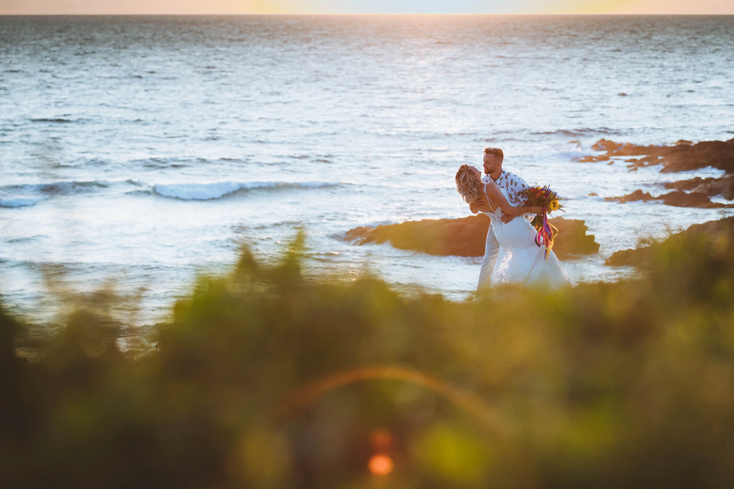 A couple in wedding attire sharing an intimate moment on a rocky beach at sunset, with the ocean in the background and blurred green foliage in the foreground.