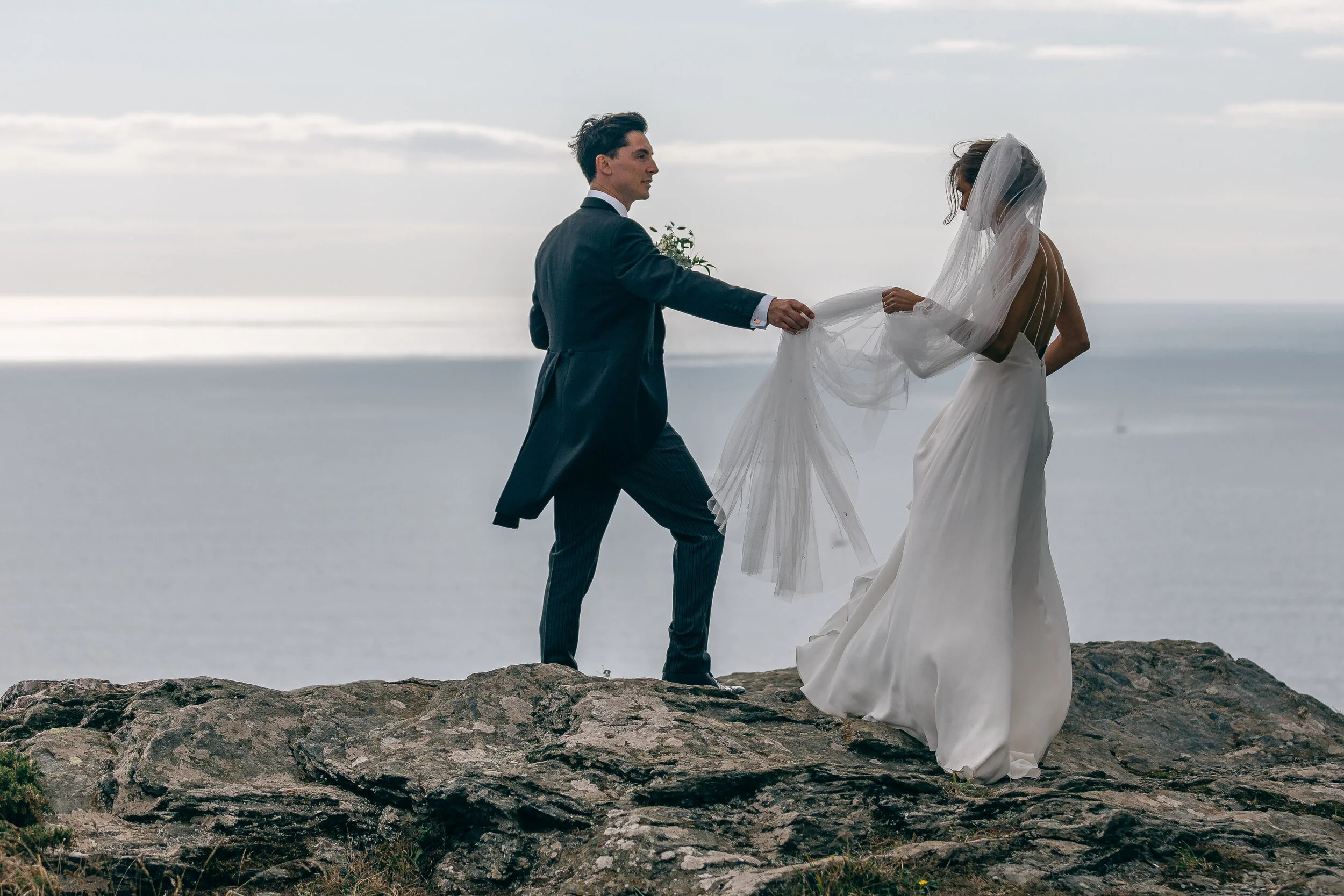 A bride in a white wedding dress with a veil and a groom in a dark suit on a rocky cliff overlooking the ocean during their wedding ceremony.