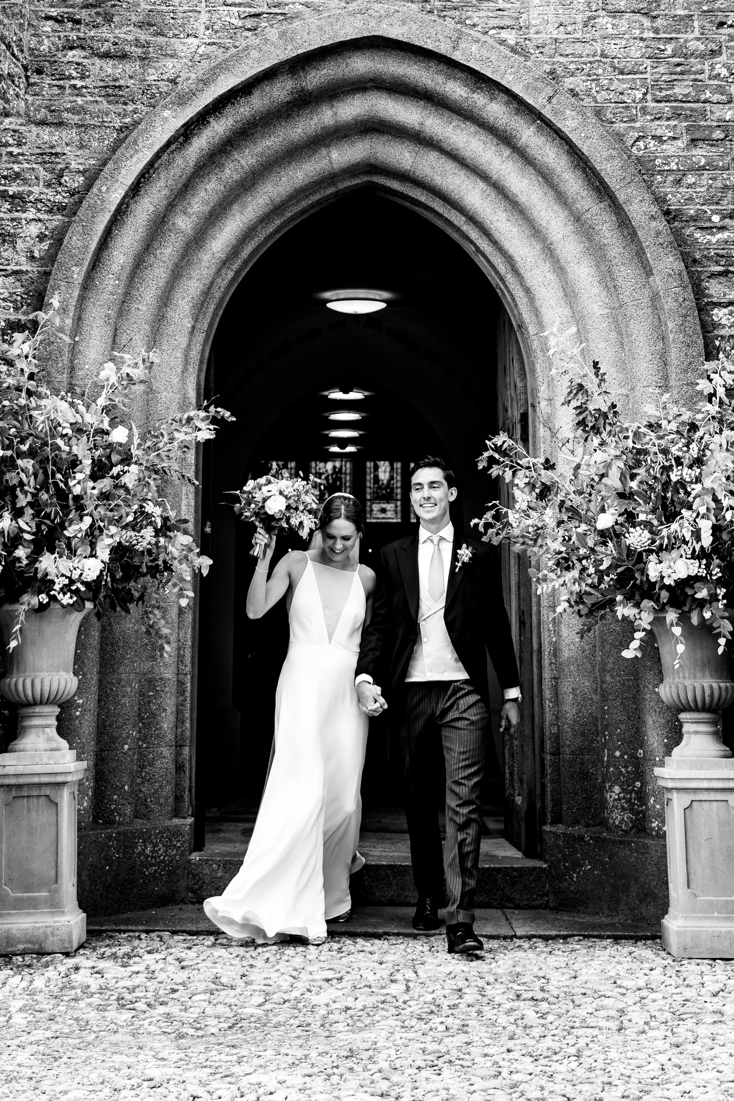 A happy bride and groom leaving a church, holding hands, with the bride holding a bouquet, surrounded by decorative plants, in black and white.
