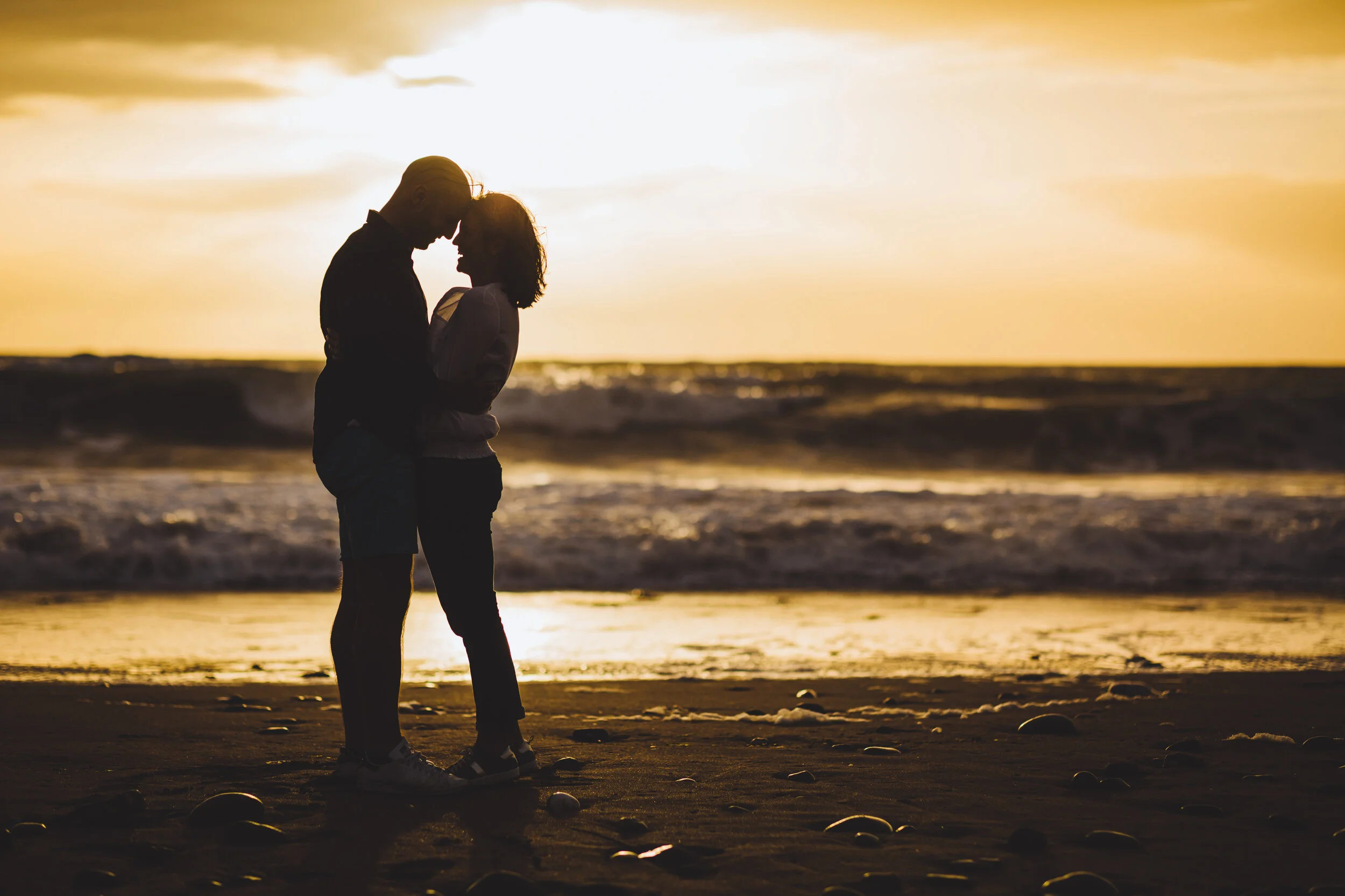 A couple kissing on Perranporth beach in Cornwall during sunset, with waves in the background and scattered rocks on the sand.