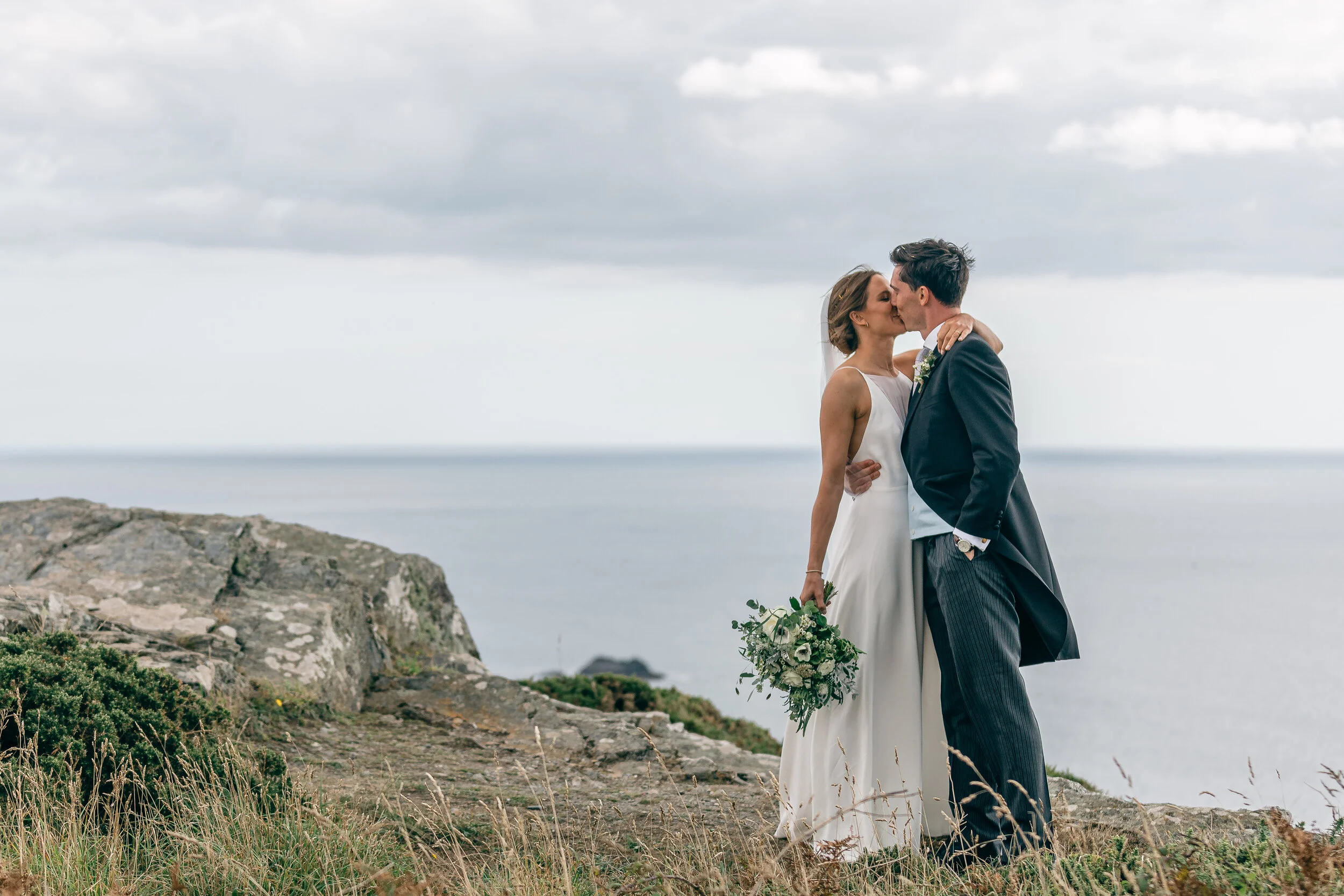 A newlywed couple sharing a kiss outdoors on a rocky cliff with a backdrop of the ocean and cloudy sky.