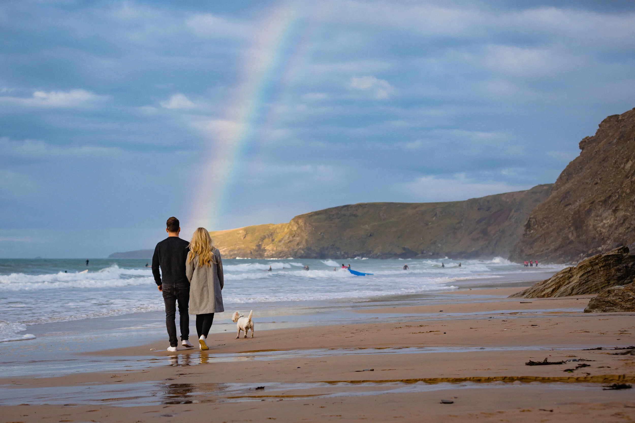 A couple walking on the beach with their dog, waves crashing nearby, a rainbow in the sky, and cliffs in the background.