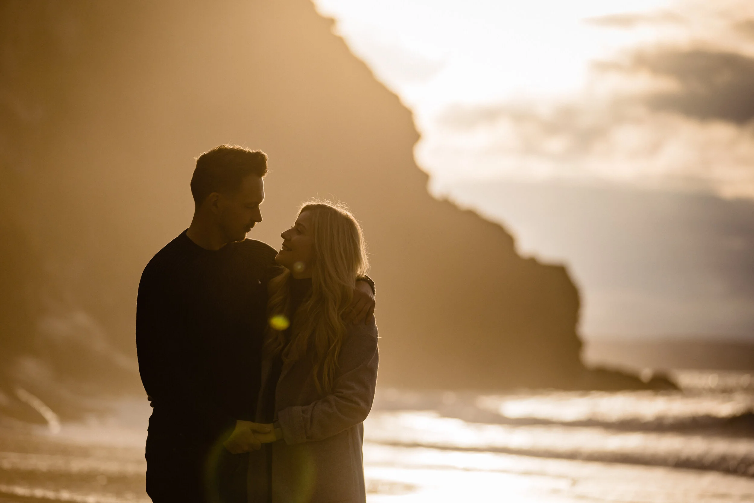 A couple standing close together on a beach during sunset, with cliffs in the background.
