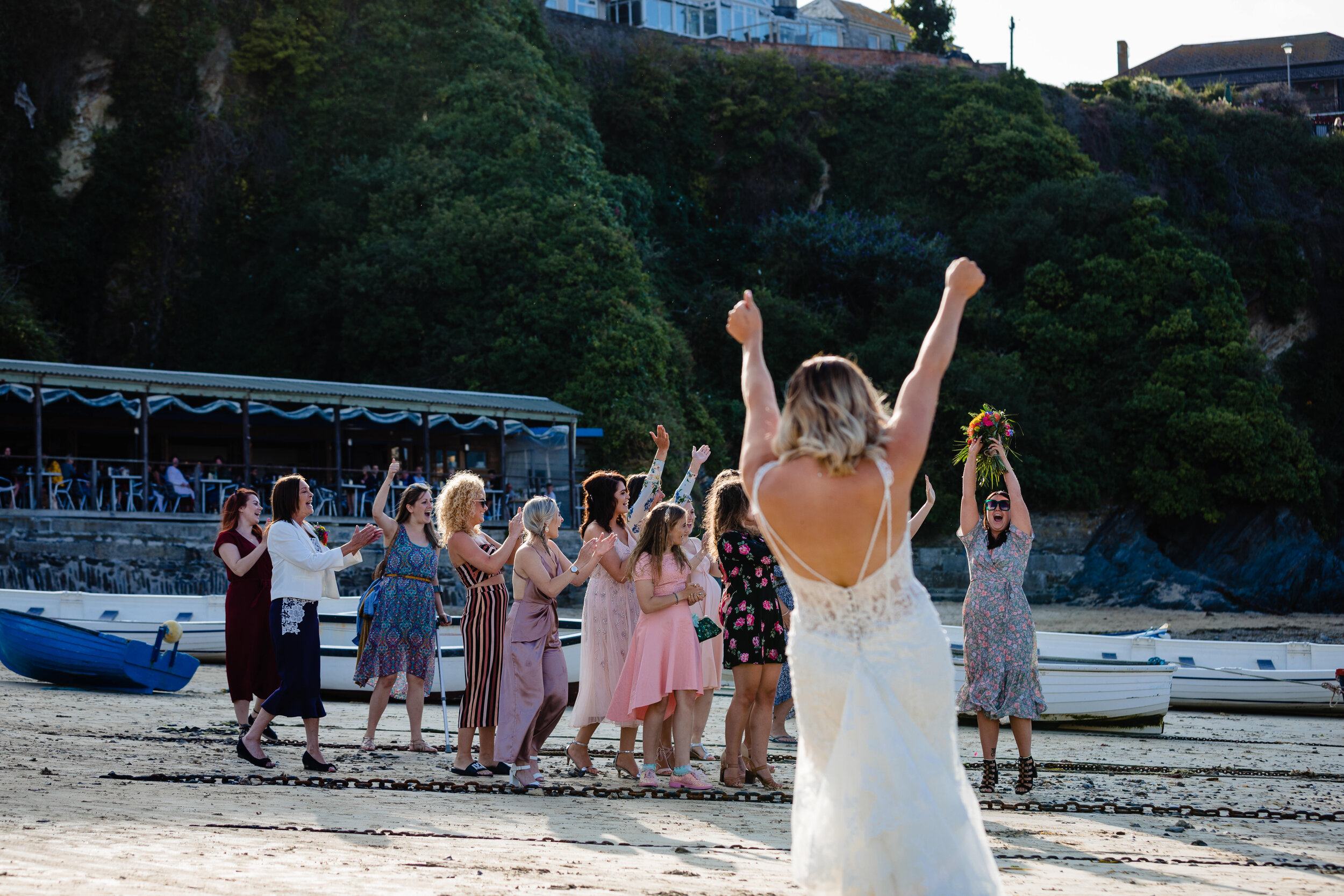 A bride in a white wedding dress stands with her back to the camera, raising her arms in celebration. A group of women on a beach in front of boats cheer, clap, and raise their hands, celebrating the wedding in Newquay, Cornwall.
