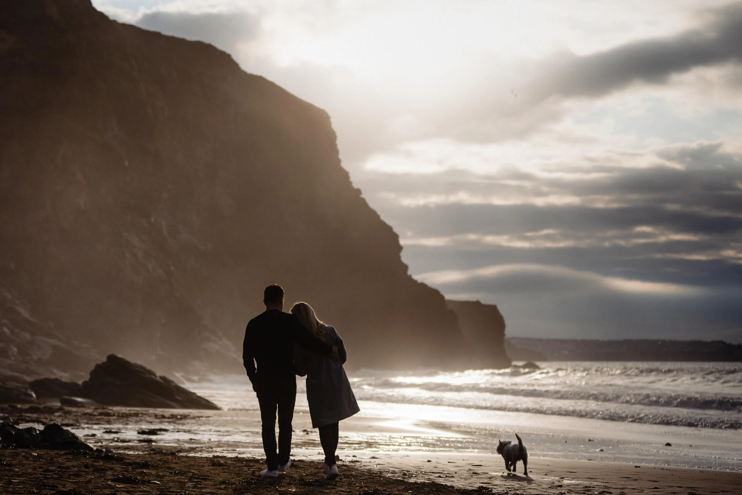 A couple walking on a beach in Cornwall with a small dog, near cliffs during sunset or sunrise.