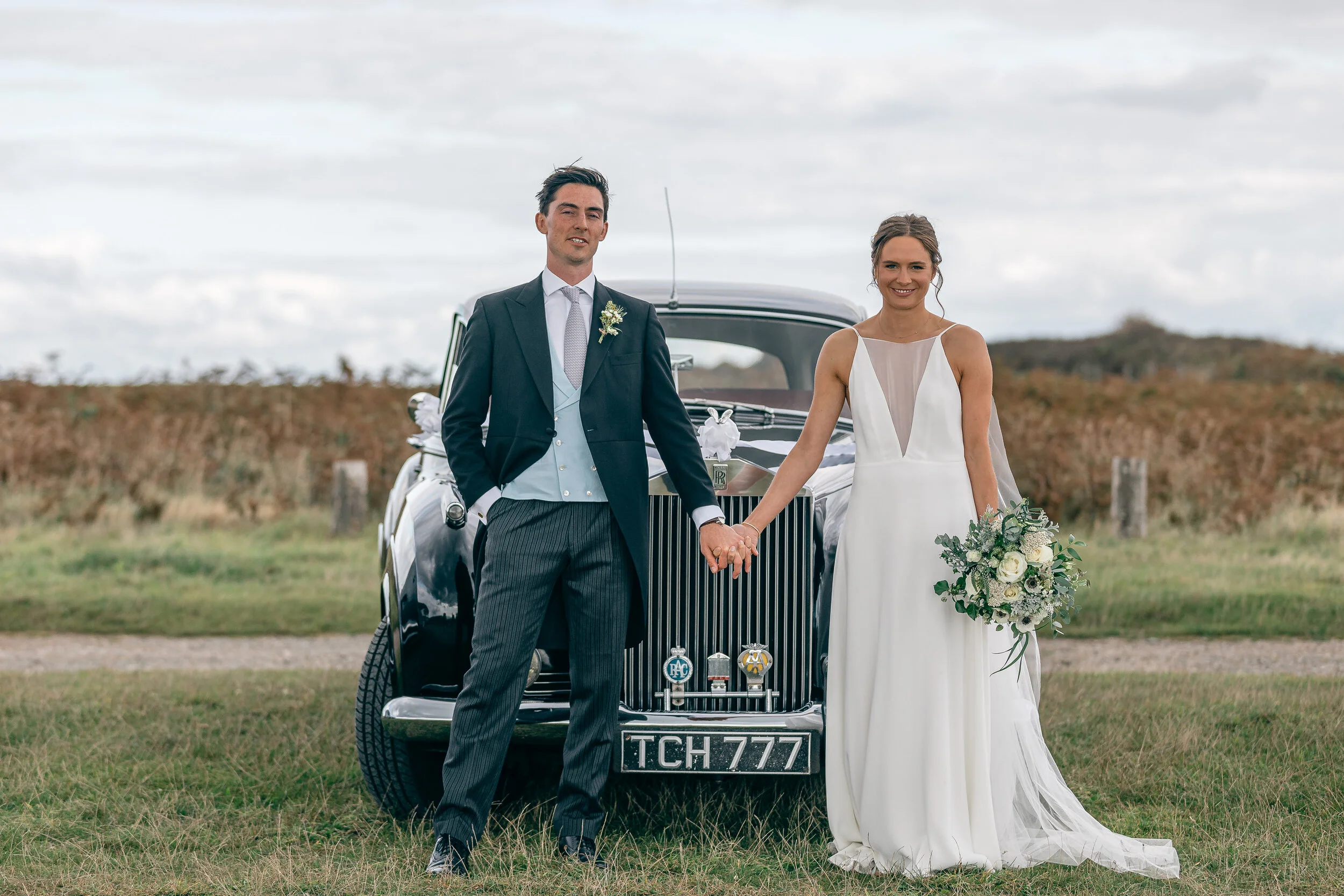 A bride and groom holding hands in front of a vintage black car with a countryside background. The bride is wearing a white dress and holding a bouquet, and the groom is in a formal suit with a boutonniere.