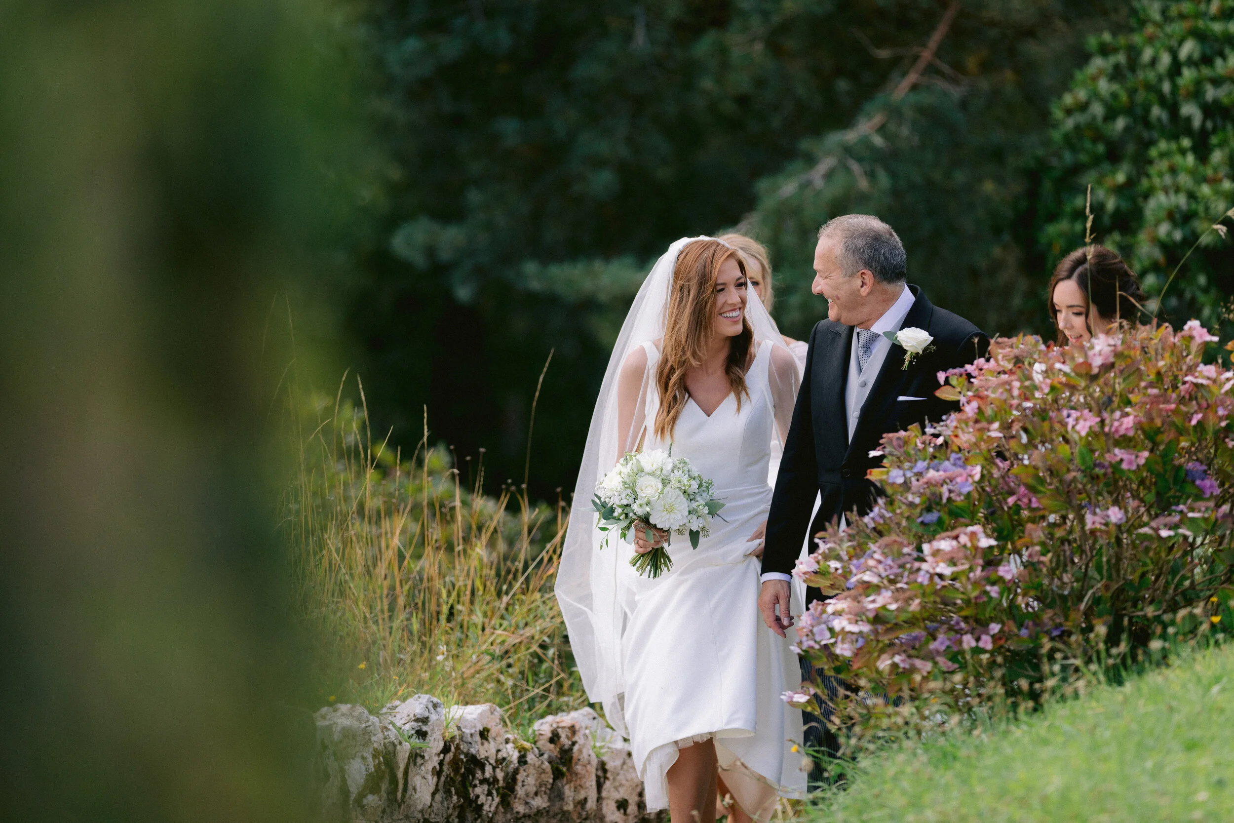 A bride in a white wedding dress and veil, holding a bouquet of white flowers, walking outdoors with an older man in a black suit, white shirt, and gray tie; they are smiling at each other.