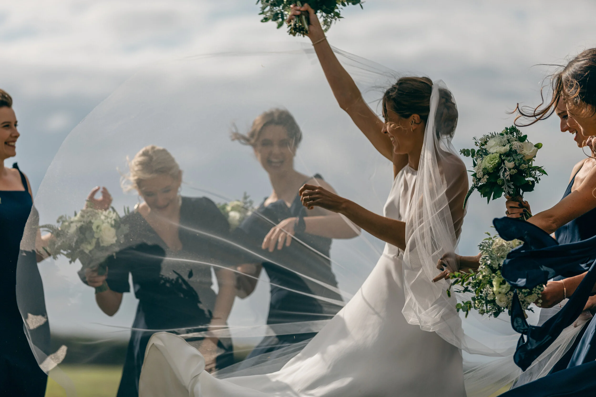 Bride in white dress and veil tossing her wedding veil in the air while surrounded by bridesmaids in navy blue dresses holding bouquets, outdoors with a cloudy sky.