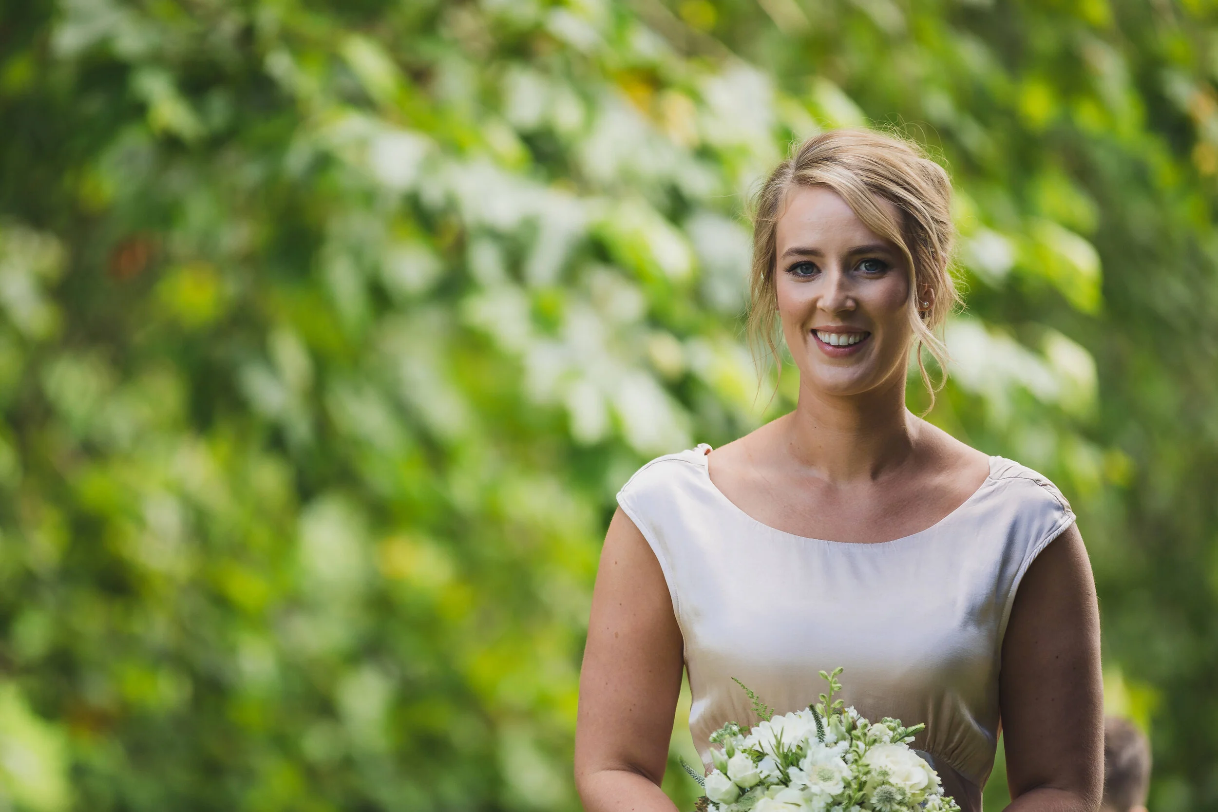 A woman with blonde hair in an updo, smiling, holding a bouquet of white flowers, outdoors near green trees.