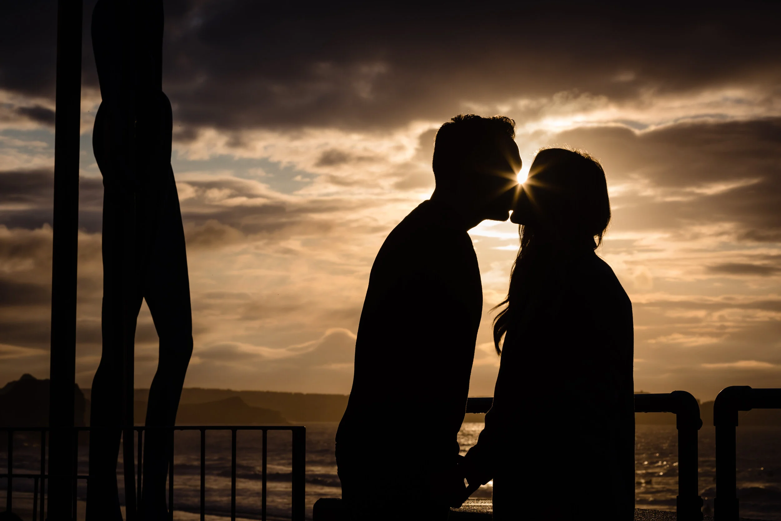 Silhouette of a couple kissing during sunset over the water, with clouds in the sky and a structure with a statue on the left.