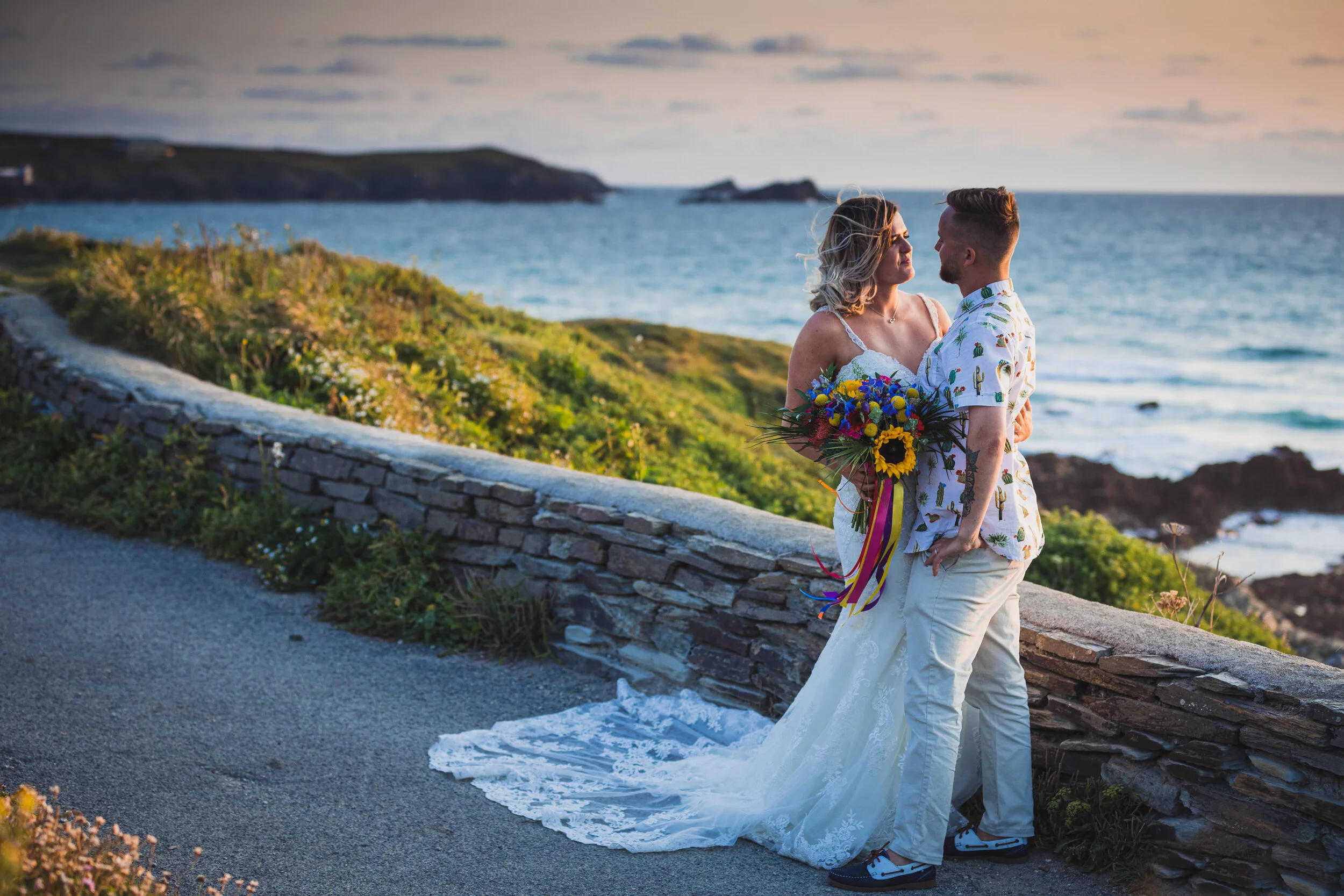 A couple in wedding attire standing by a stone wall near the ocean at sunset, with the woman holding a colorful bouquet.