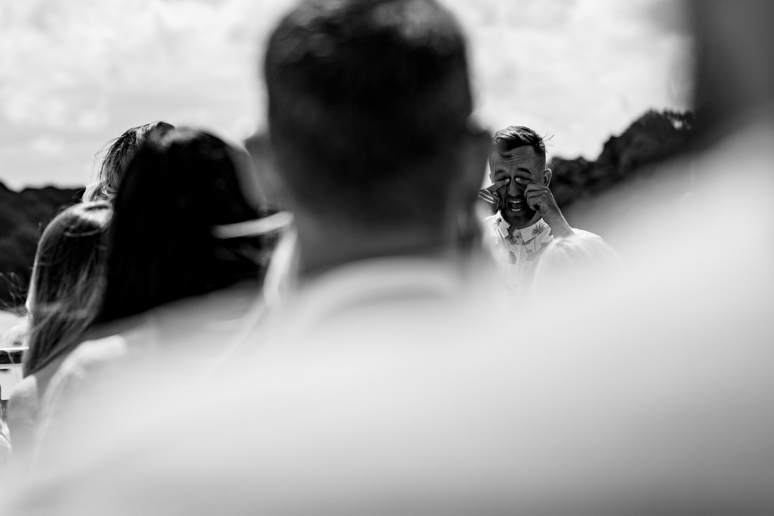 Man wiping tears while standing outdoors, surrounded by people, with landscape in the background.