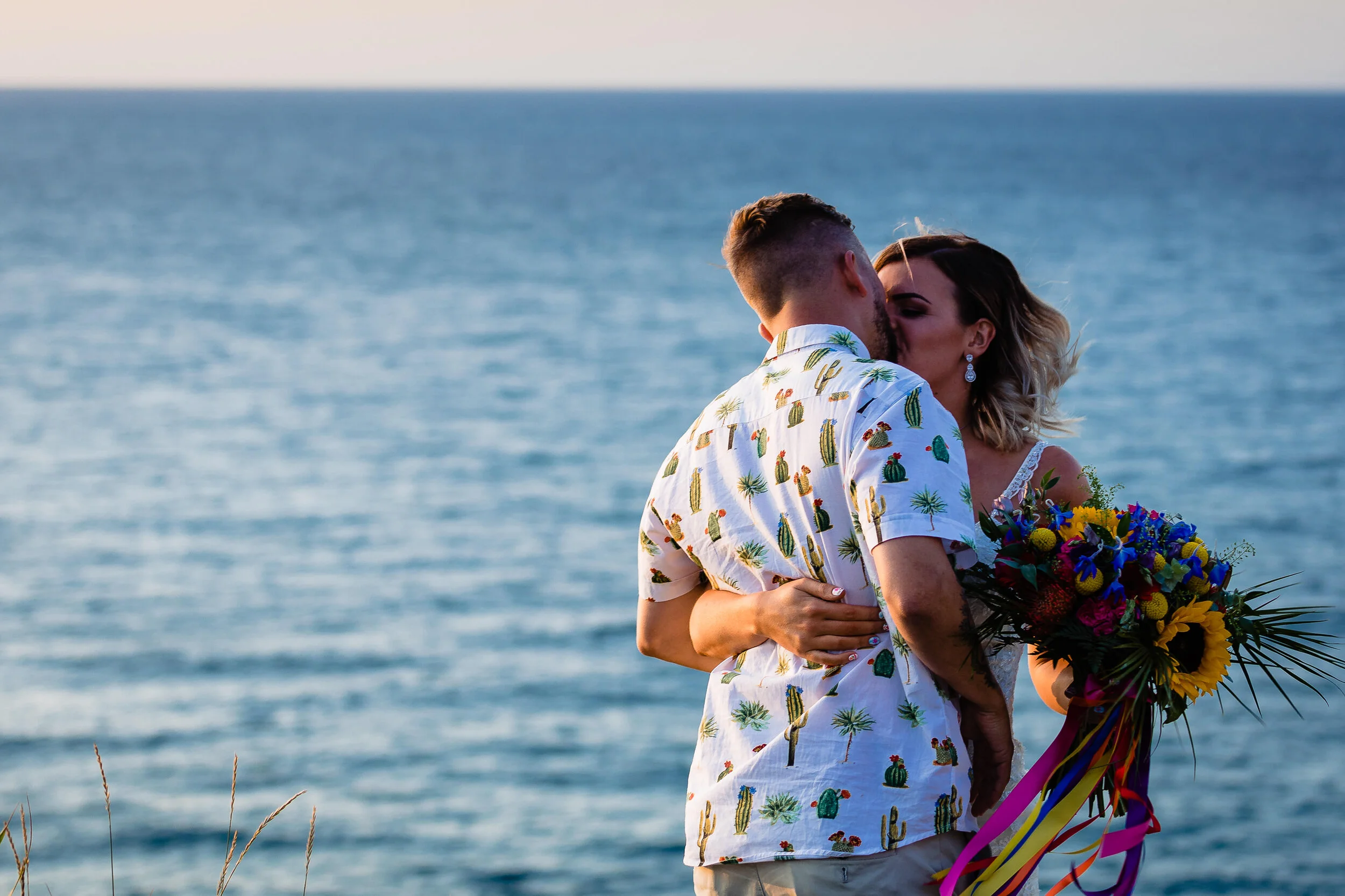 A couple kissing by the water, the woman holding a colorful bouquet of flowers.