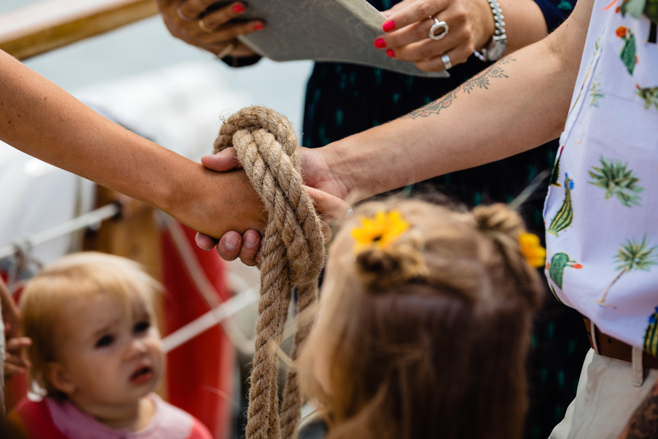 Two people shaking hands over a thick rope, with children and a person holding a clipboard in the background, at an outdoor event.
