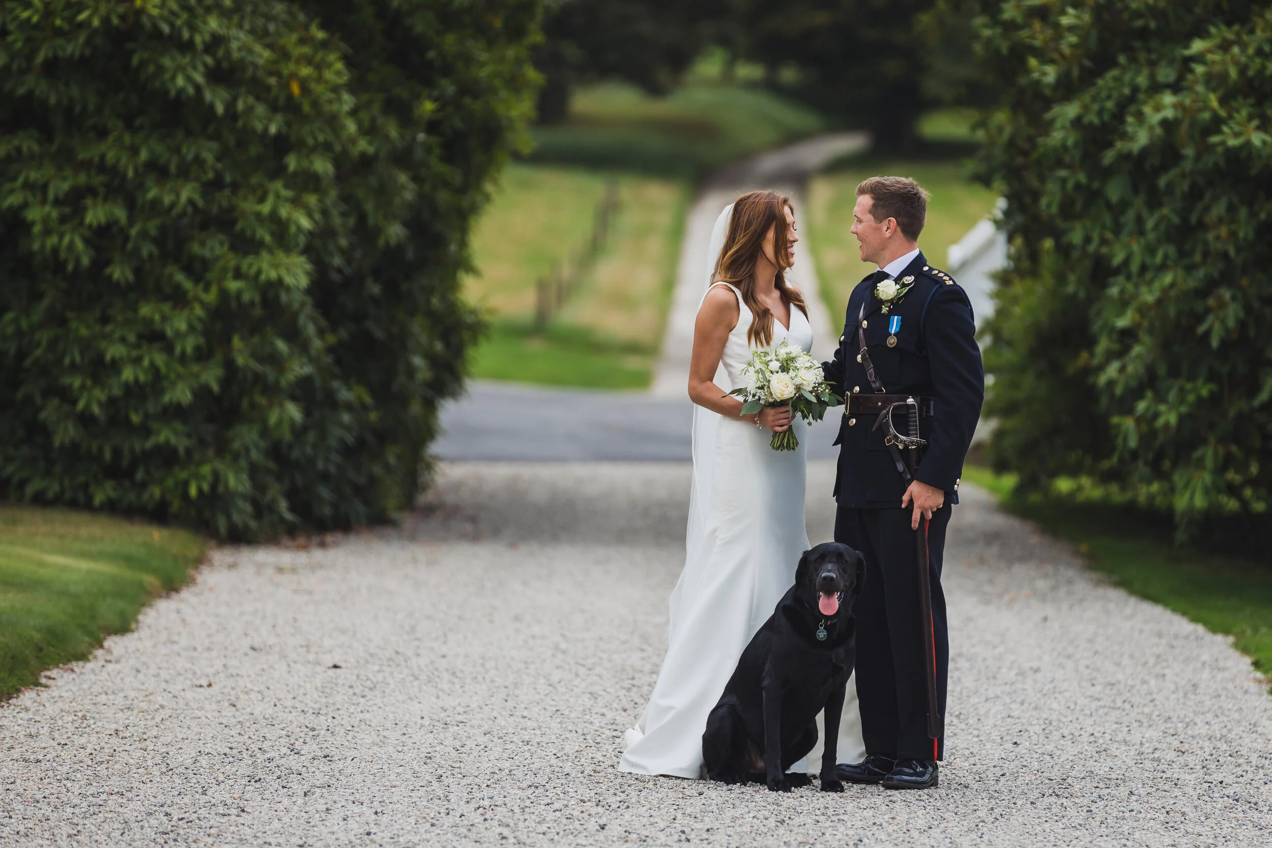Bride and groom, a soldier in uniform, standing on a gravel path with a black dog, surrounded by greenery, during a wedding celebration.
