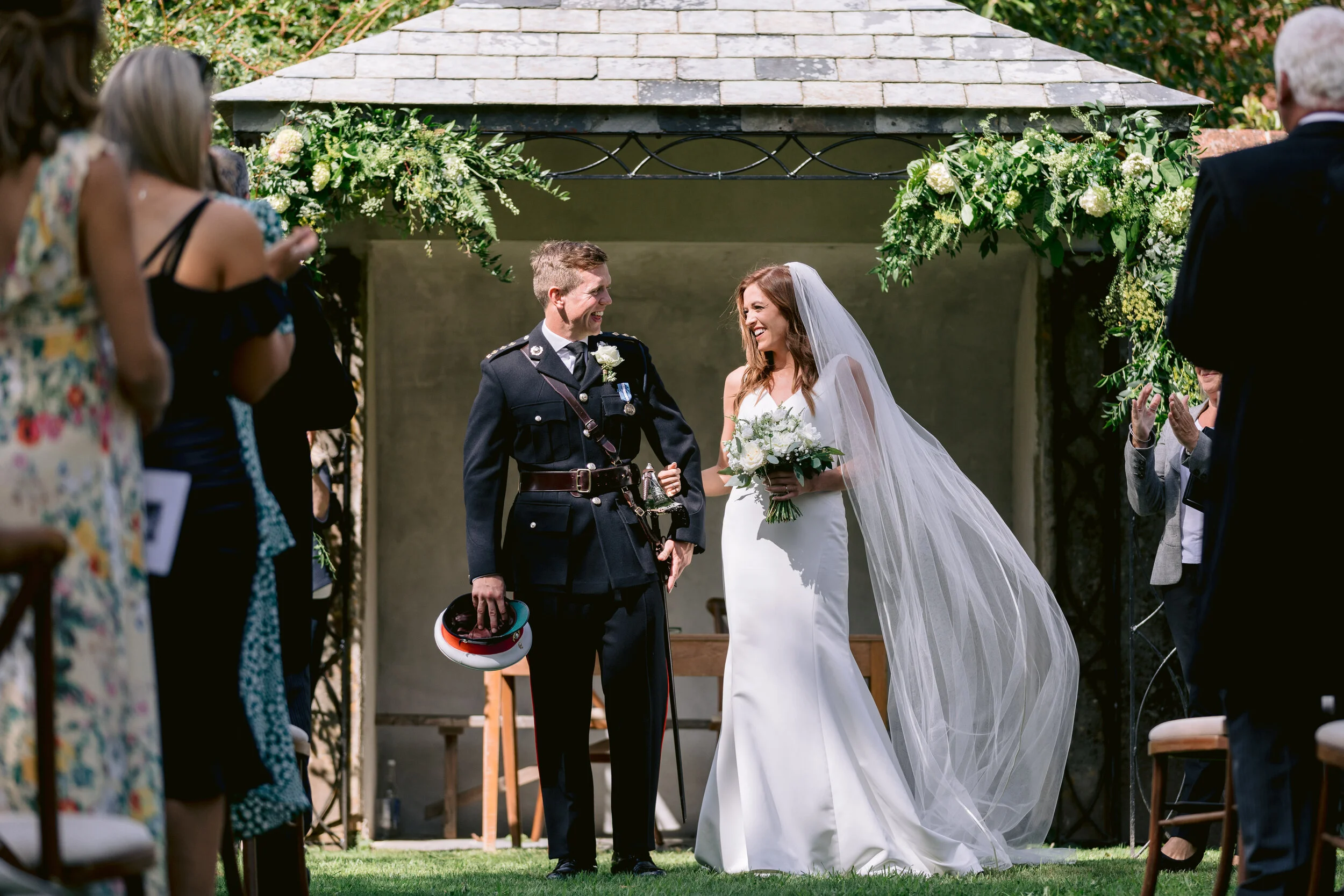 A bride and groom sharing a joyful moment during their outdoor wedding ceremony, surrounded by friends and family. The bride wears a white wedding dress and veil, and holds a bouquet of white flowers. The groom is in military uniform, holding his hat