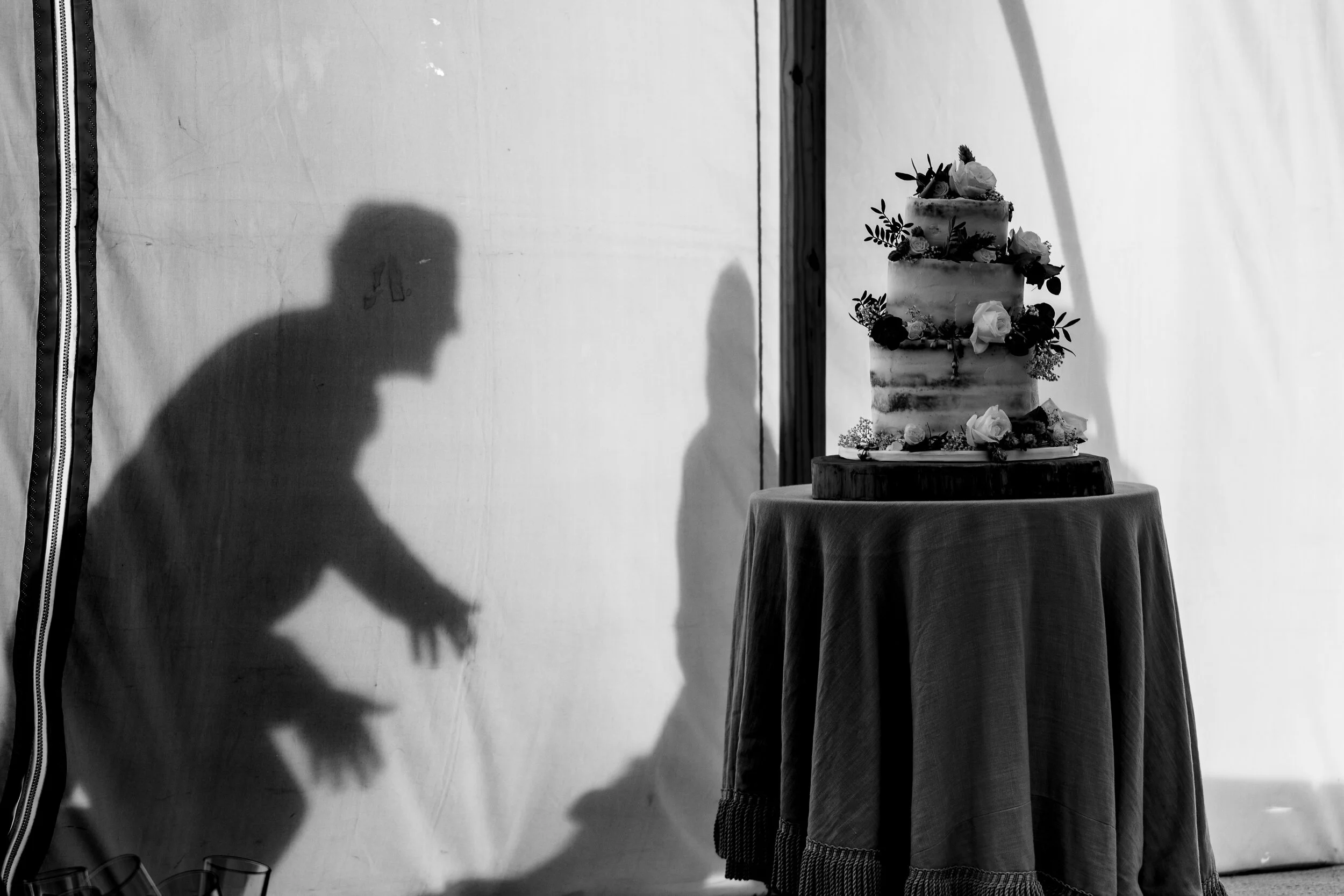 Shadow of a person reaching towards a four-tier wedding cake decorated with flowers on a table.