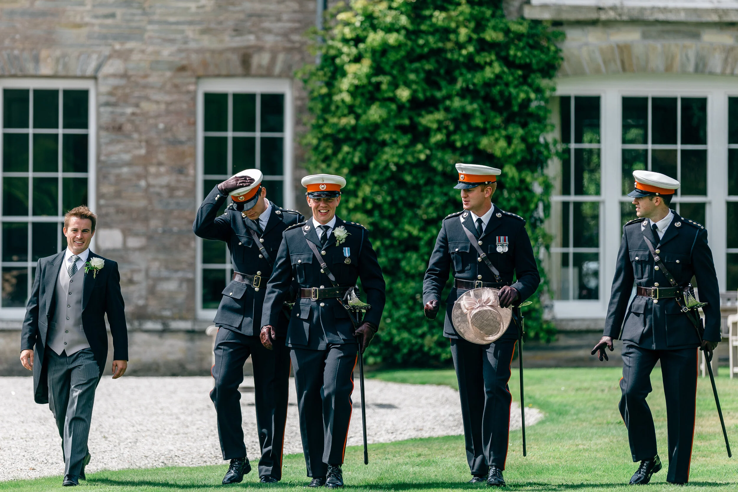 A groom in a gray suit and four police officers in uniform walking on a lawn in front of a stone building with large windows and green ivy. The police officers are dressed in black formal uniforms with hats, and one salutes while the others smile and