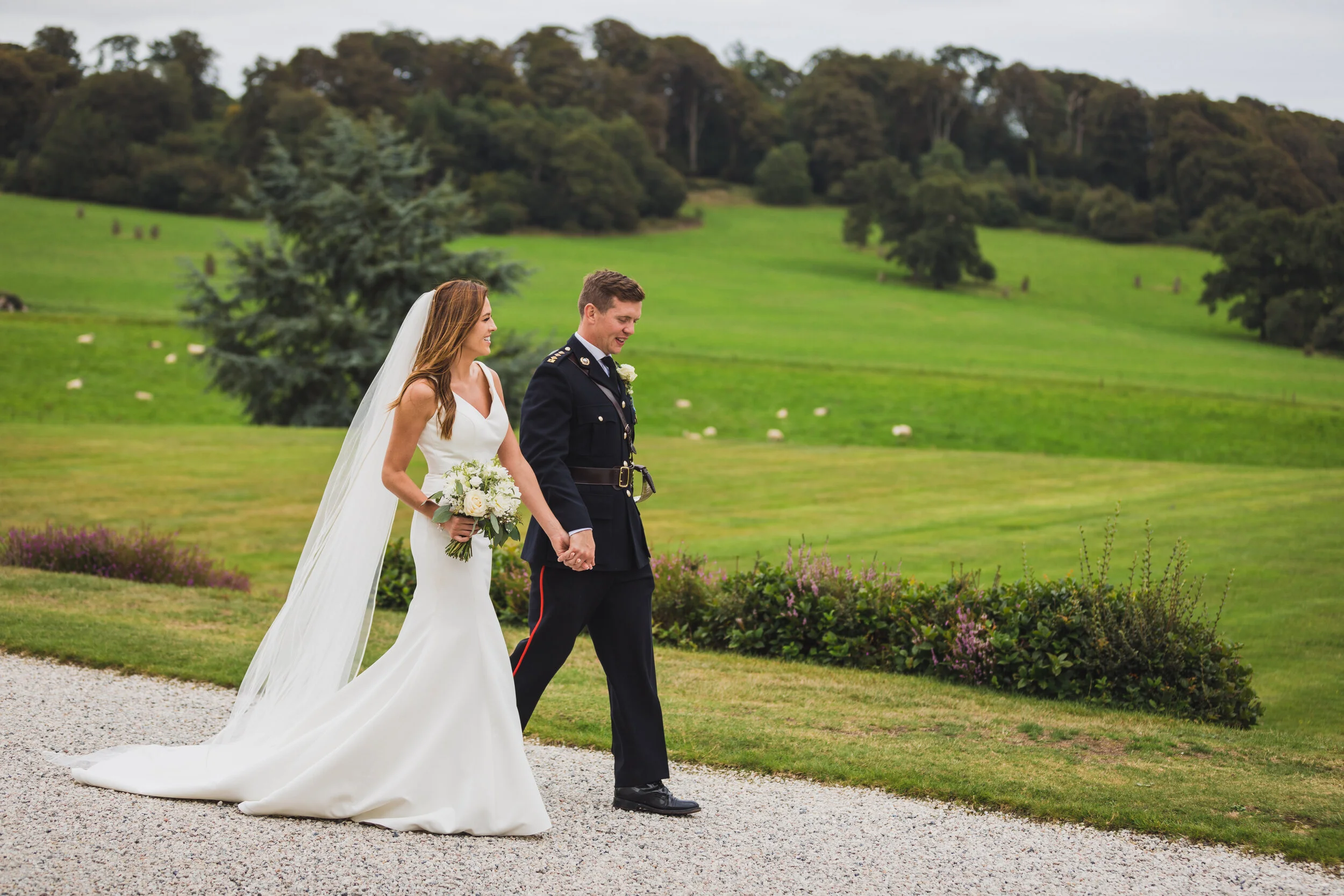 A bride and groom walking hand in hand outdoors on a gravel path with a lush green landscape and trees in the background.