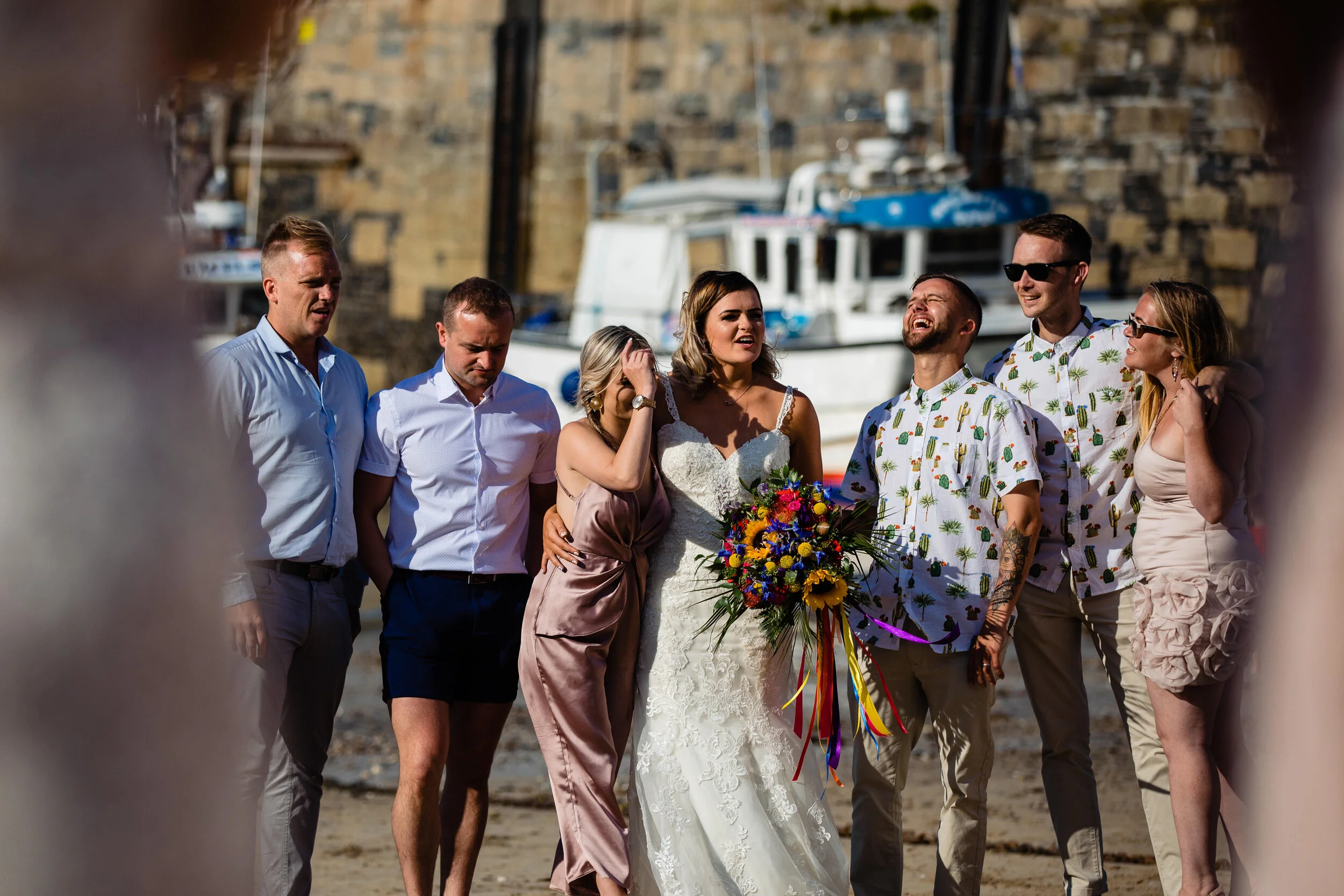 A group of seven people, including a bride in a white wedding dress holding a colorful bouquet, standing on a beach with boats and a stone wall in the background, enjoying a moment together.