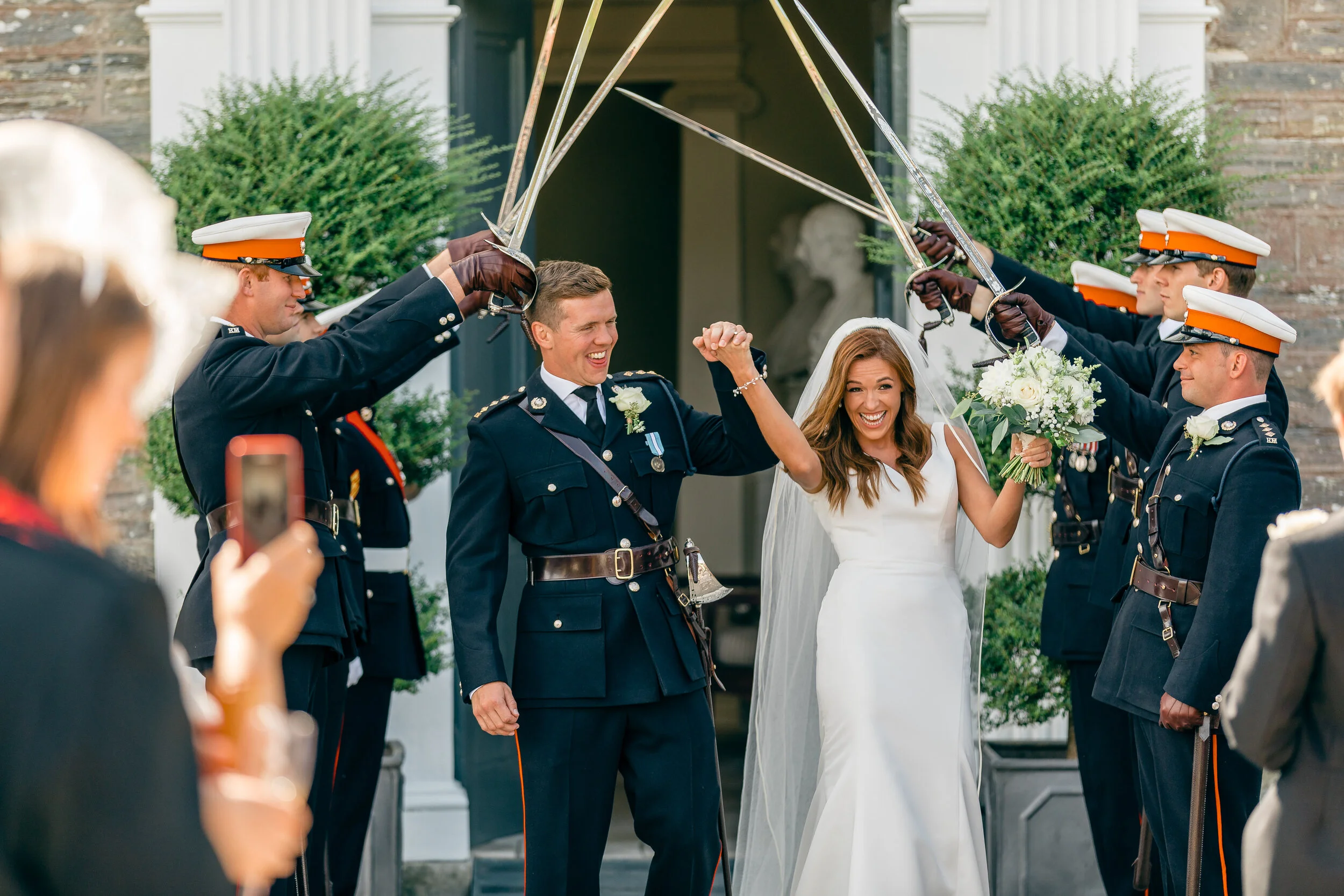 Bride and groom celebrating as military honor guards create an arch with swords during wedding ceremony outside building with large potted plants.