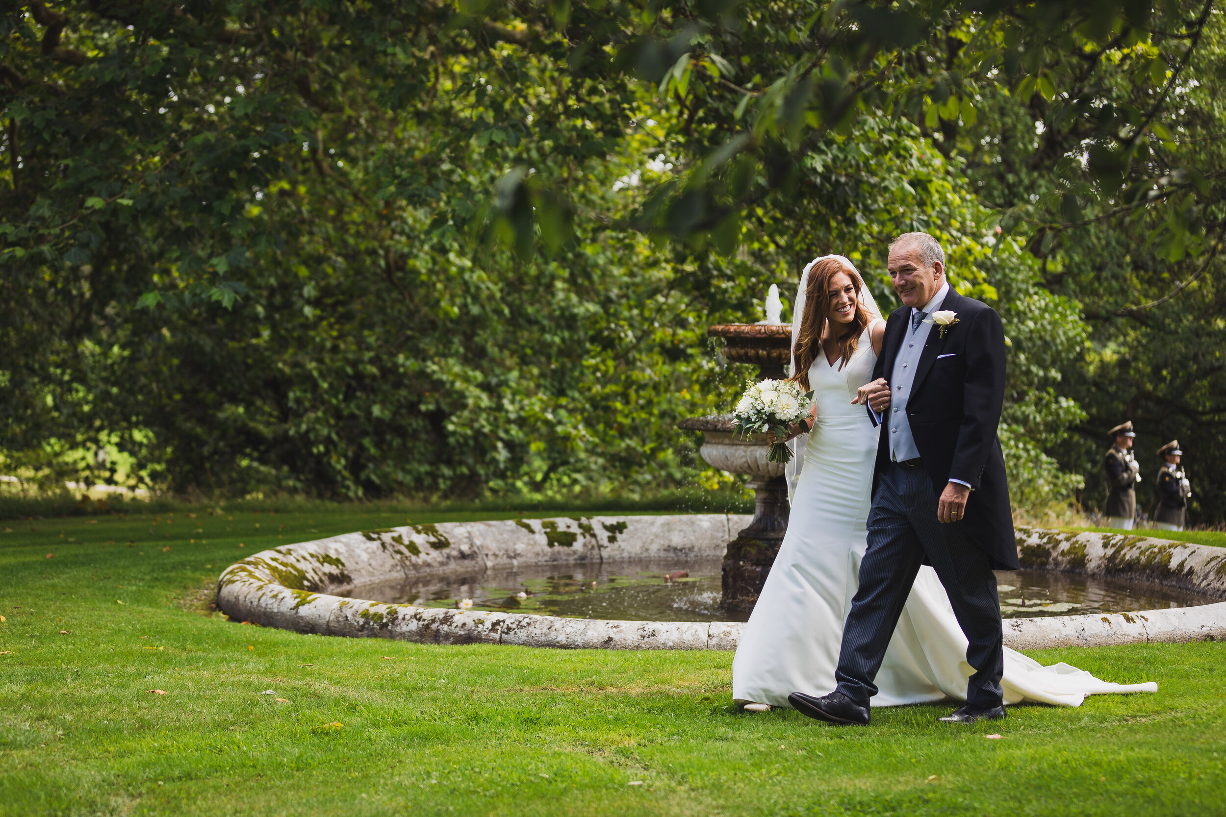 A bride and an older man, possibly her father, walking arm-in-arm in a garden with a fountain in the background during a wedding. The bride is in a white wedding dress holding a bouquet of white flowers, and the man is in a formal tuxedo with a bouto