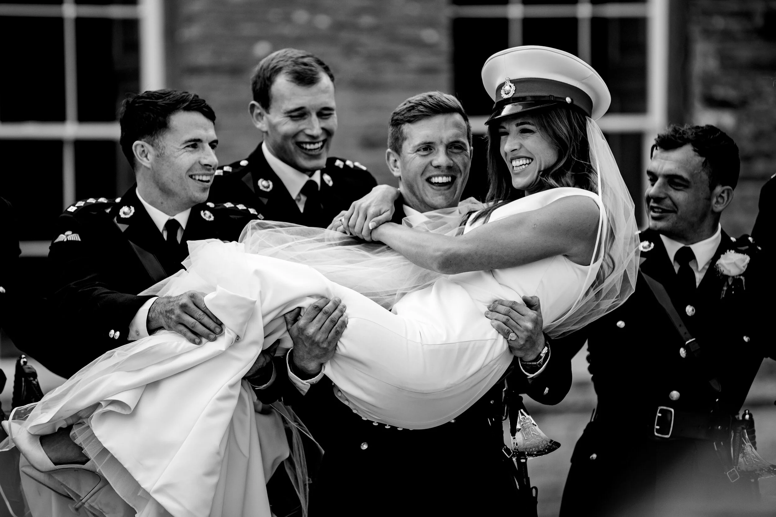 A bride in a wedding dress and uniform hat being carried by five uniformed men in military attire, smiling and laughing outdoors.