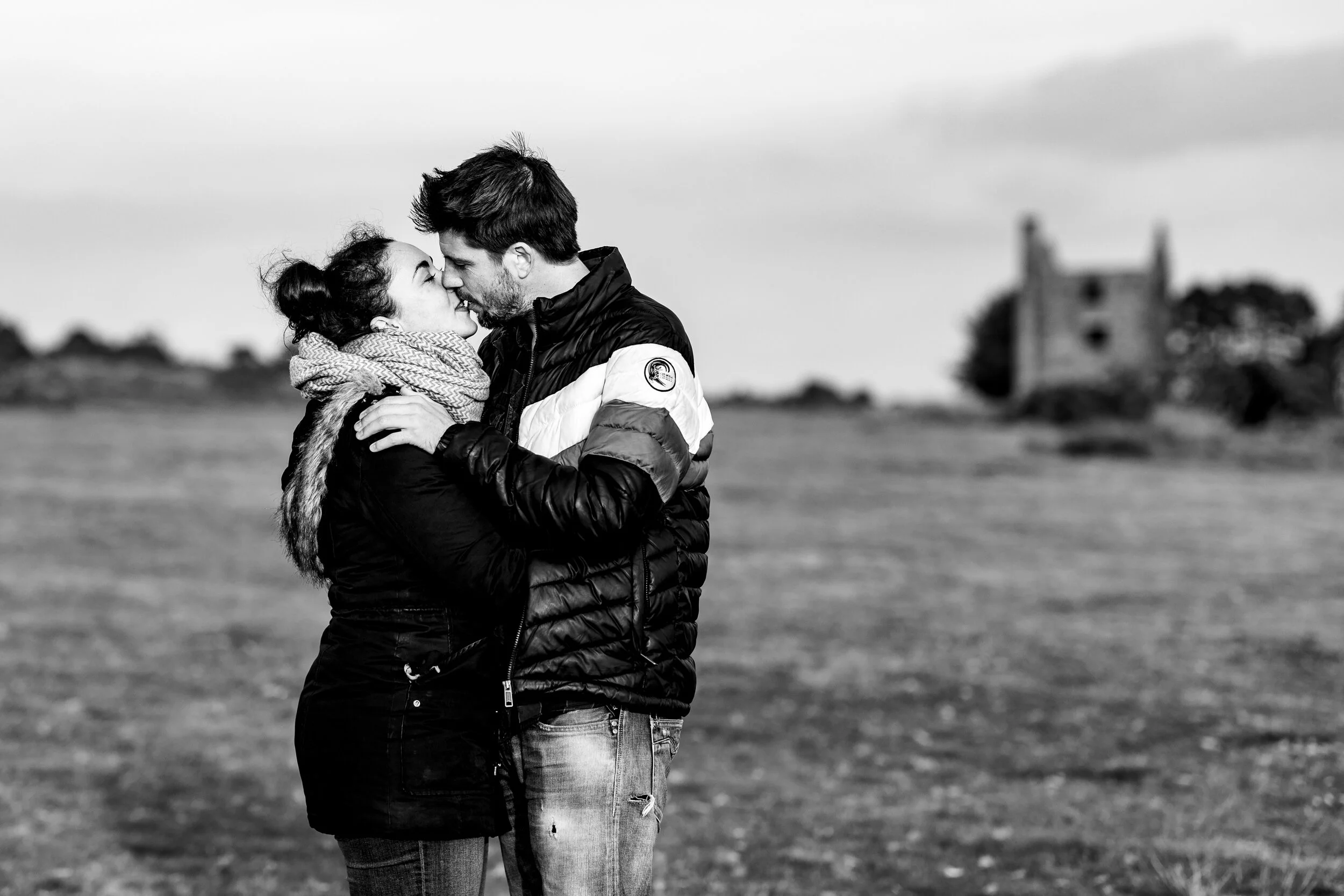 A couple embracing outdoors in Cornwall, preparing to kiss, with a blurred castle in the background, black and white photograph.