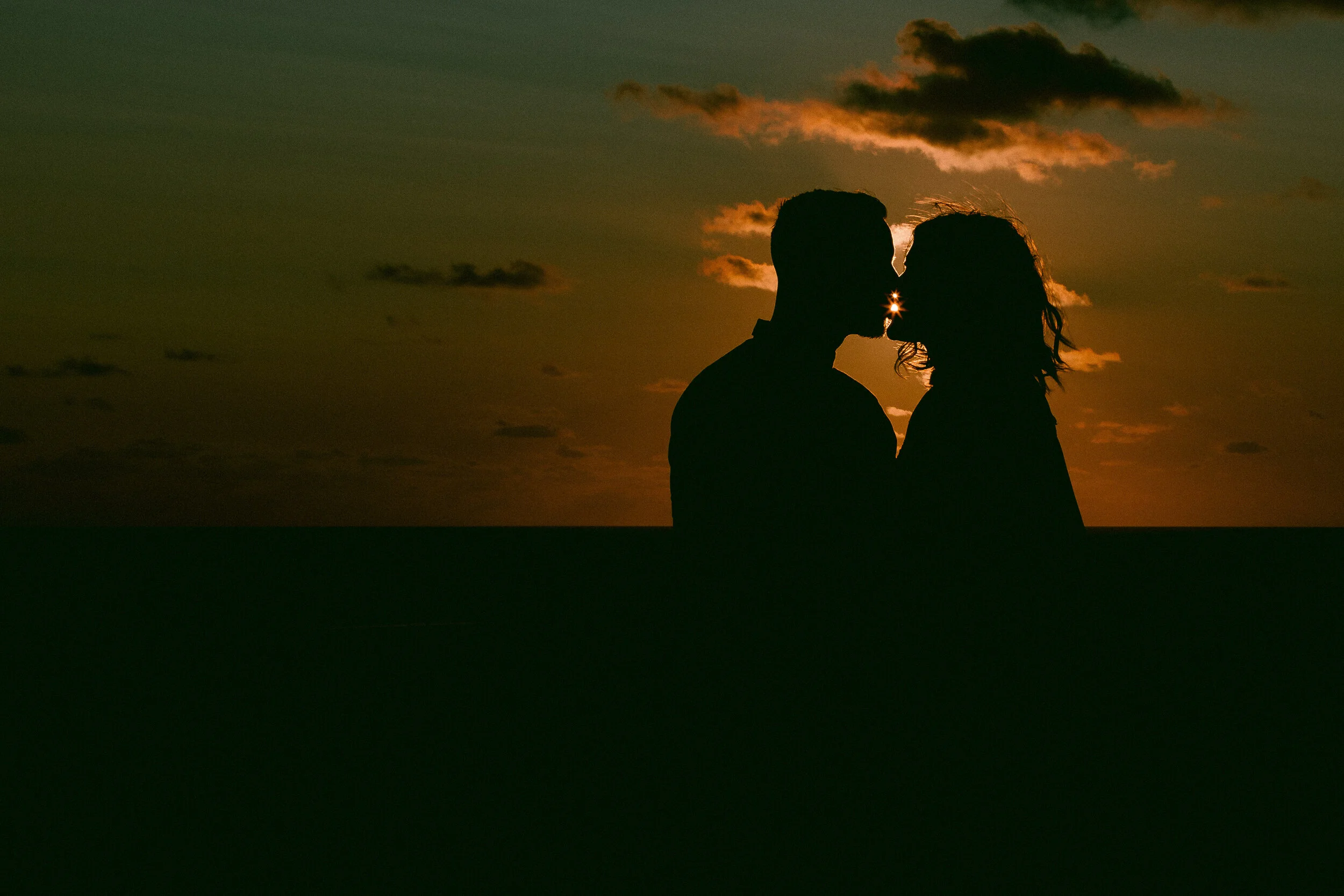 Silhouette of a couple kissing at sunset with the sun shining between them and clouds in the sky.