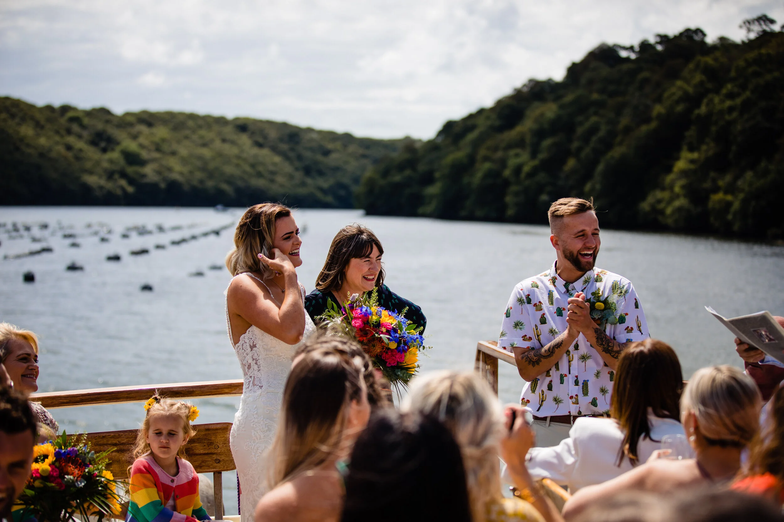 A wedding ceremony taking place outdoors on a boat with a river and green hills in the background. The bride in a white lace dress is smiling, holding her face, while the groom in a colorful short-sleeve shirt laughs. Guests are seated around them, i
