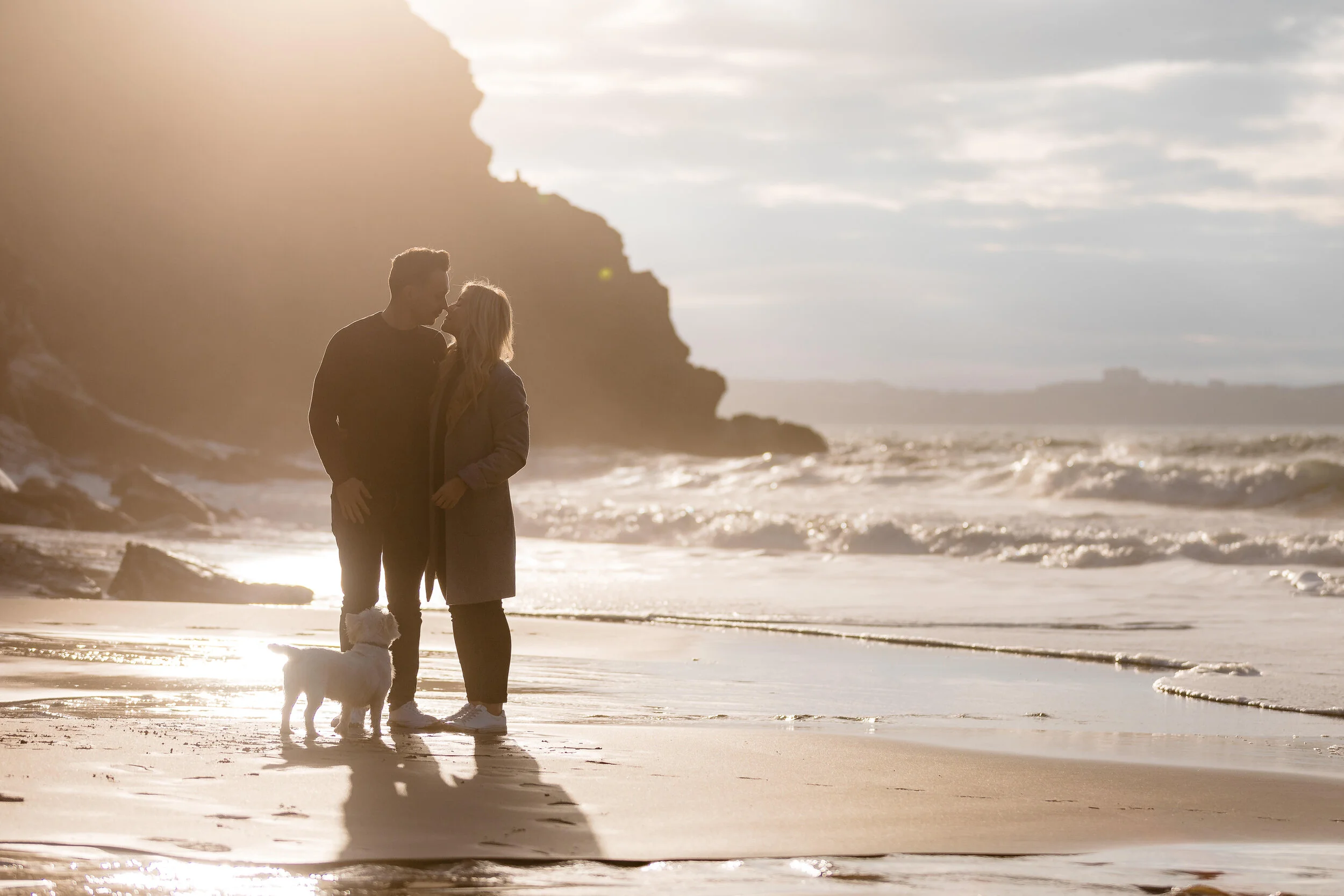 A couple and their dog standing on a beach during sunset, with waves and a rocky cliff in the background.