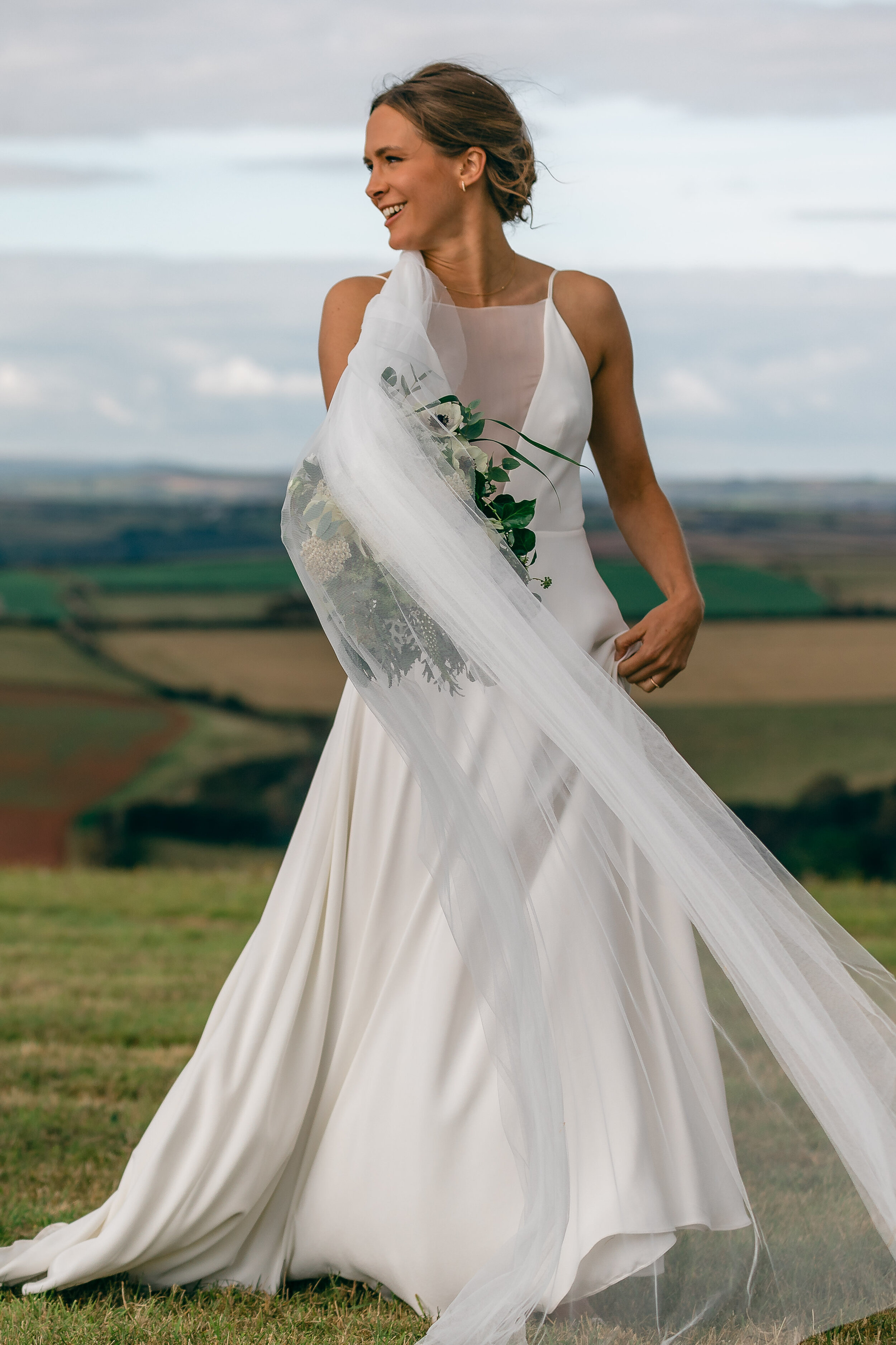 A woman in a wedding dress holding a bouquet outdoors against a landscape background.