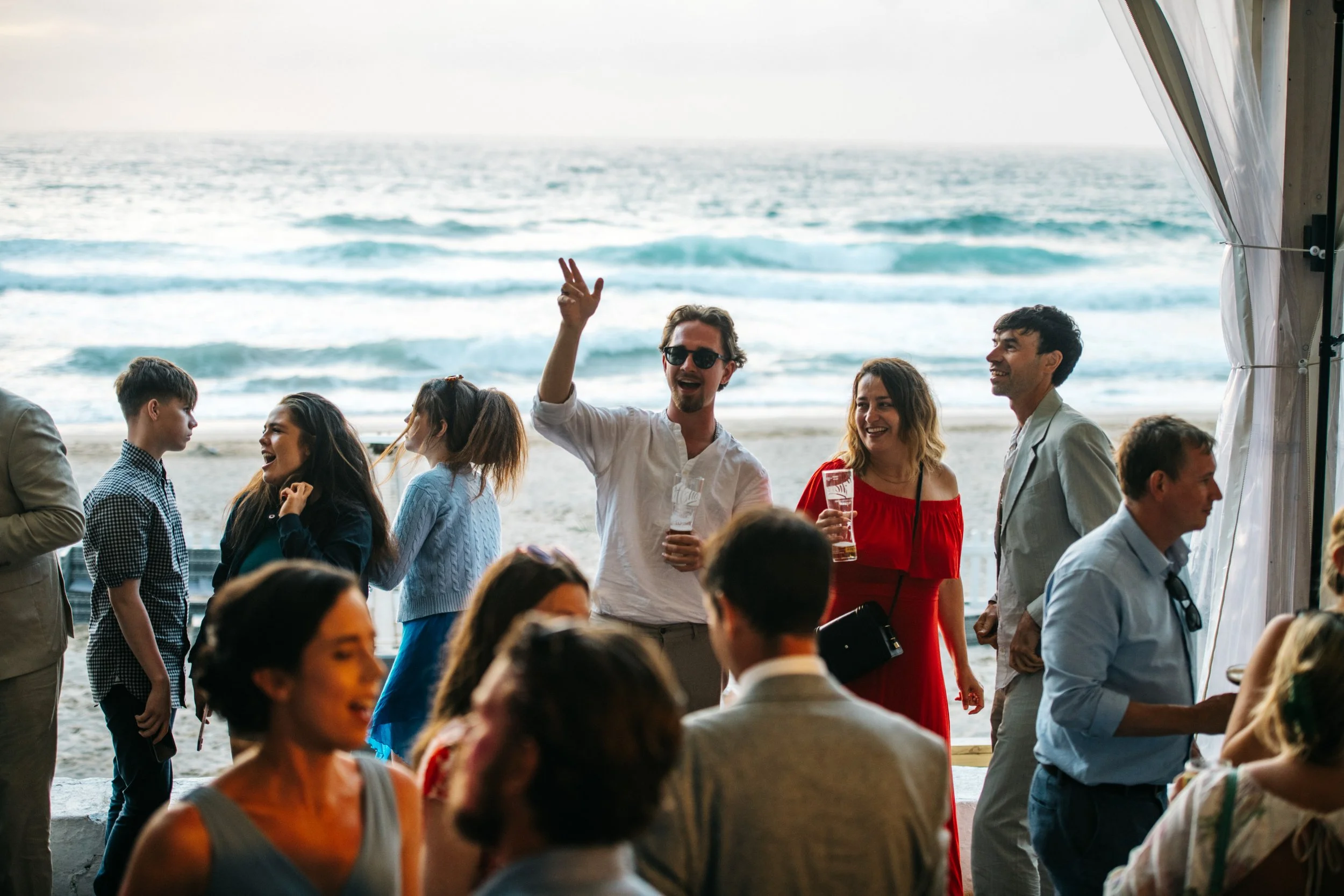 People enjoying a beachside event with the ocean in the background, some dancing, laughing, and holding drinks.
