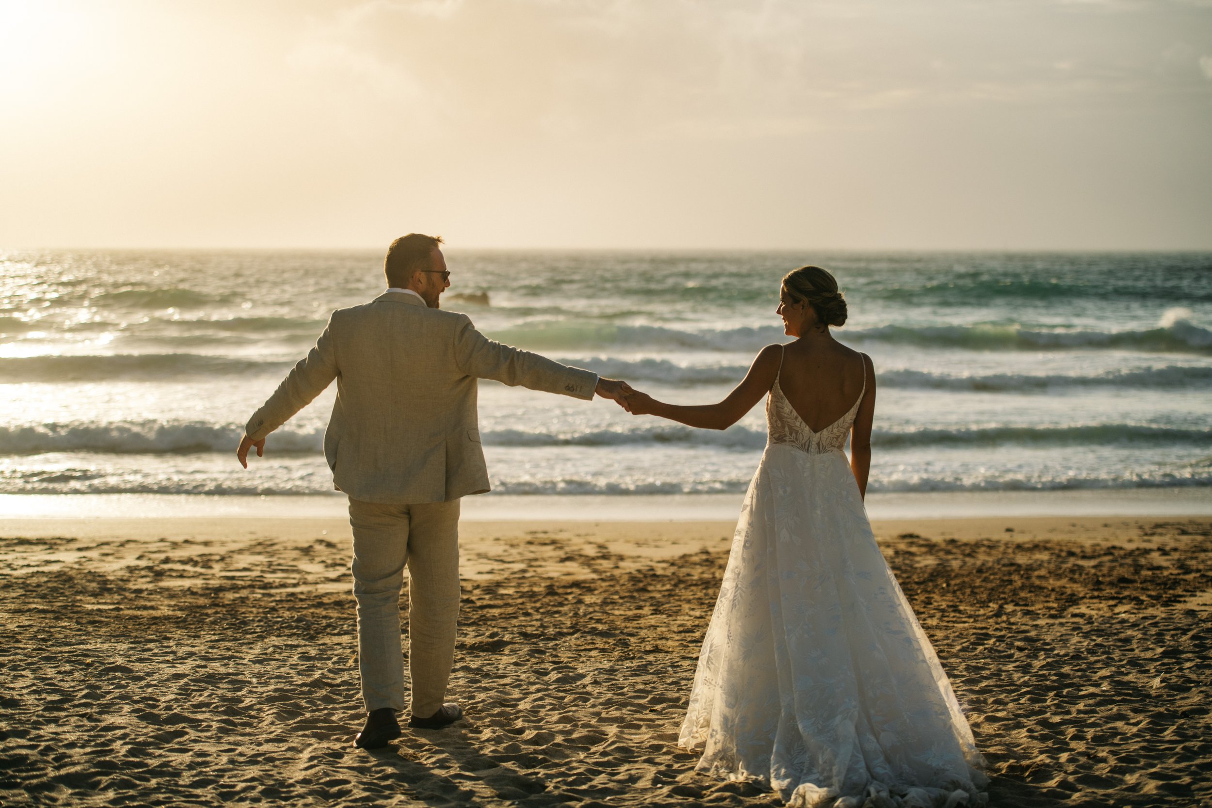 A bride and groom holding hands on a beach during sunset, with waves in the background.