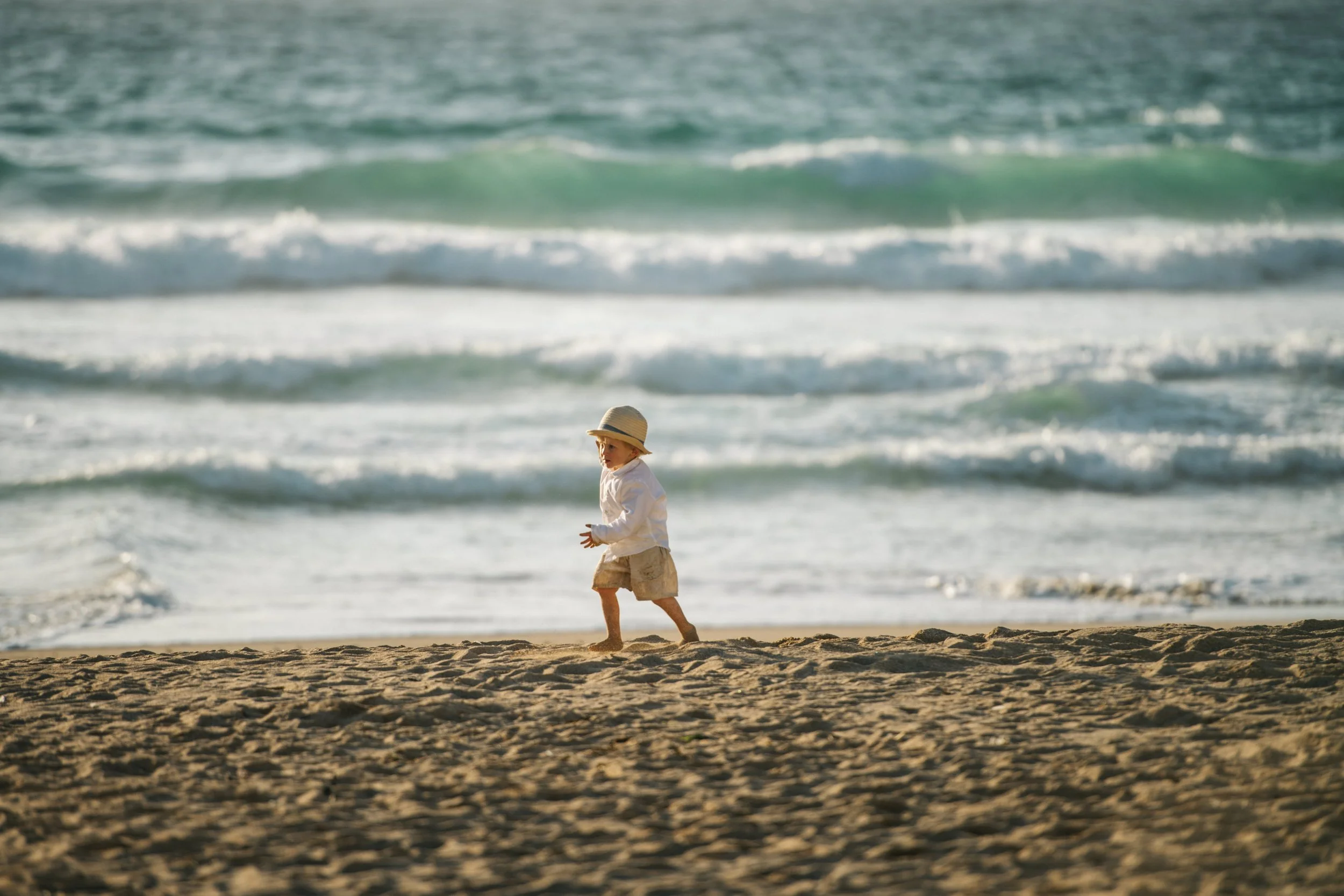 A young child wearing a white shirt, khaki shorts, and a straw hat walking on the sandy beach near the ocean with small waves in the background.