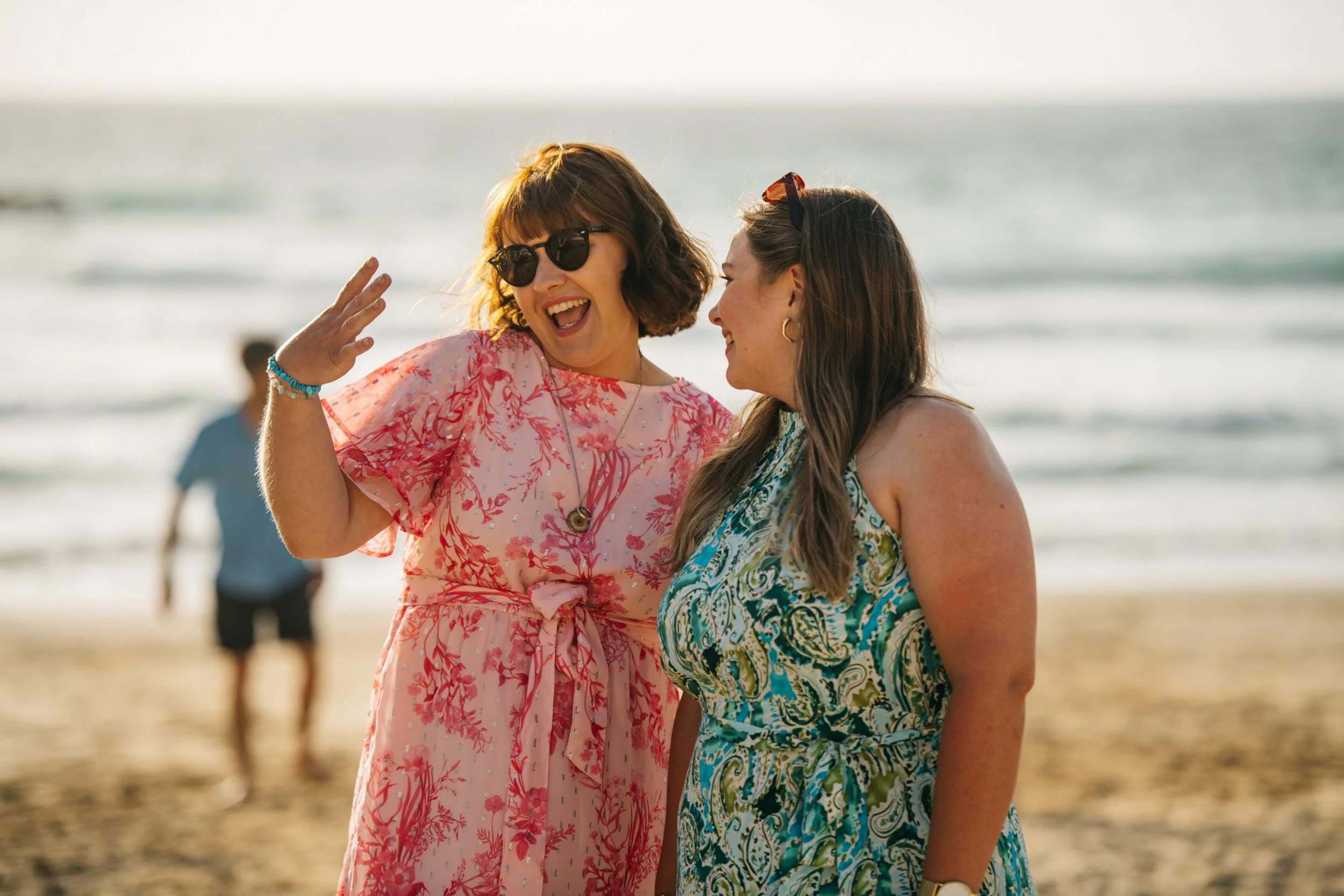 Two women are smiling and talking at the beach, with ocean waves in the background.