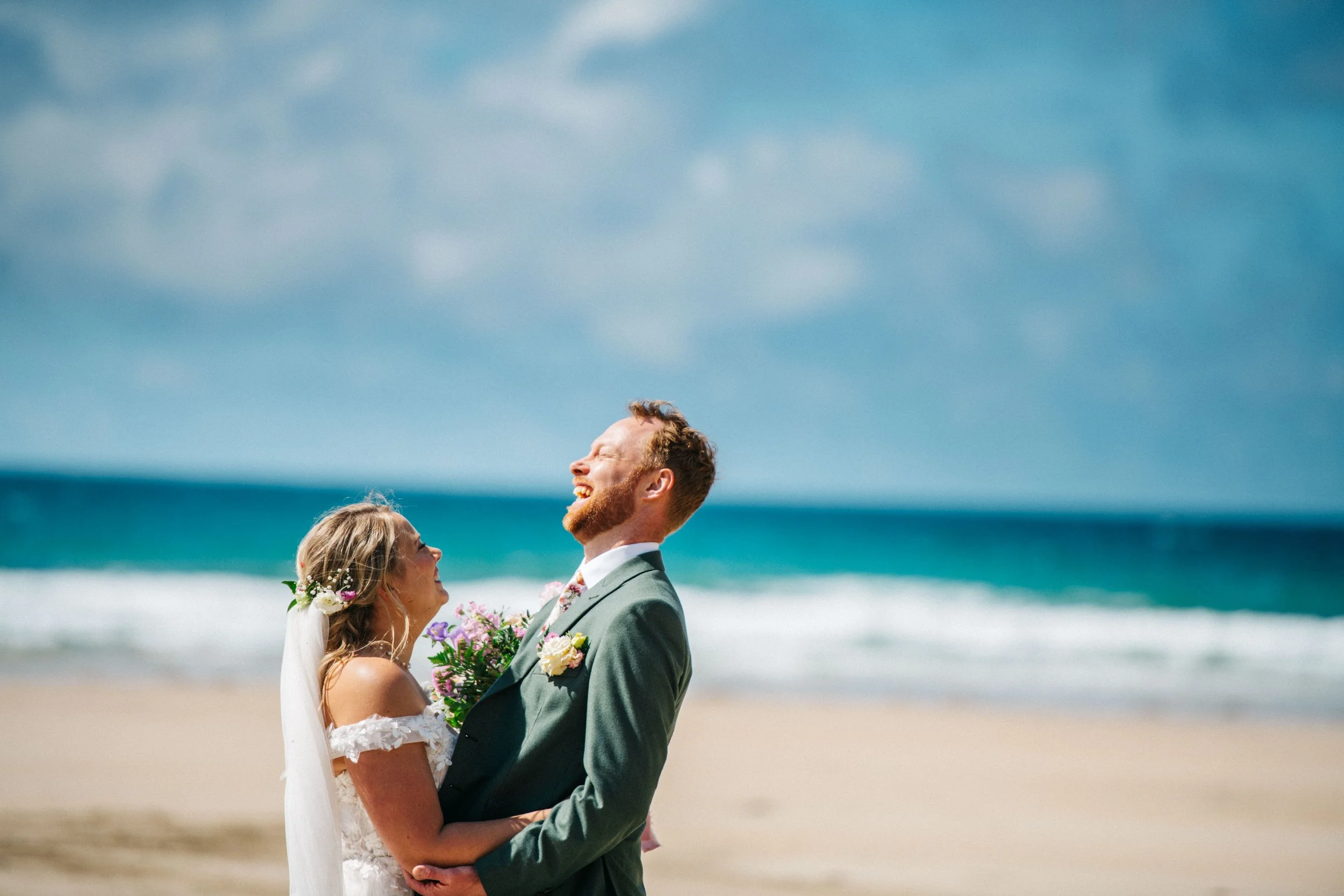 A bride and groom at the beach, smiling and holding each other, with ocean waves and a cloudy sky in the background.