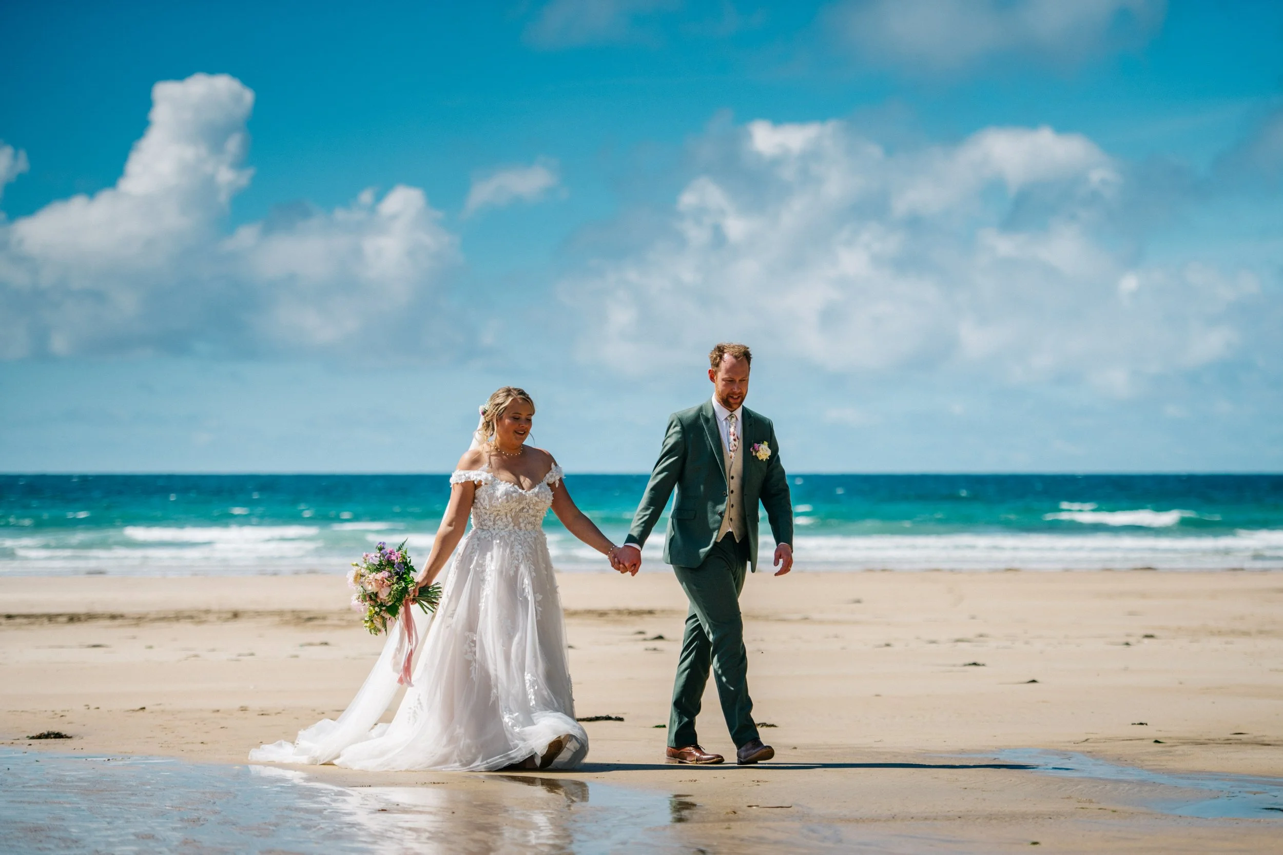 A bride and groom walking hand in hand on the beach near the ocean, with blue sky and clouds above.