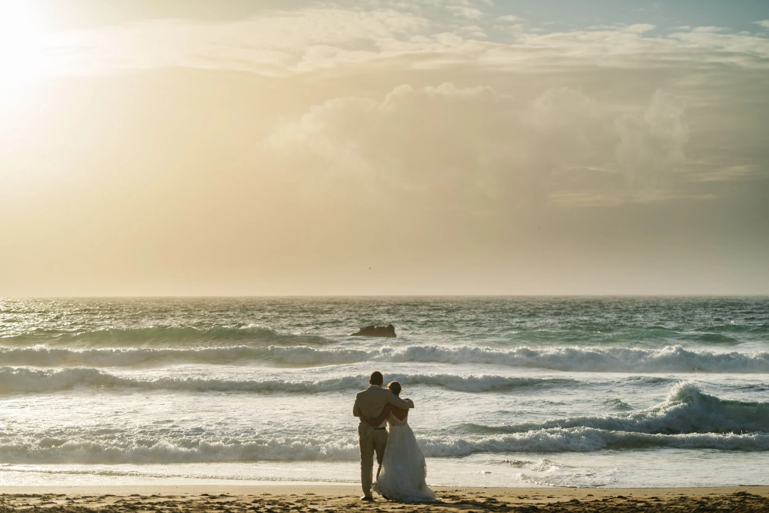 A couple standing close together at the beach during sunset, facing the ocean with waves crashing.