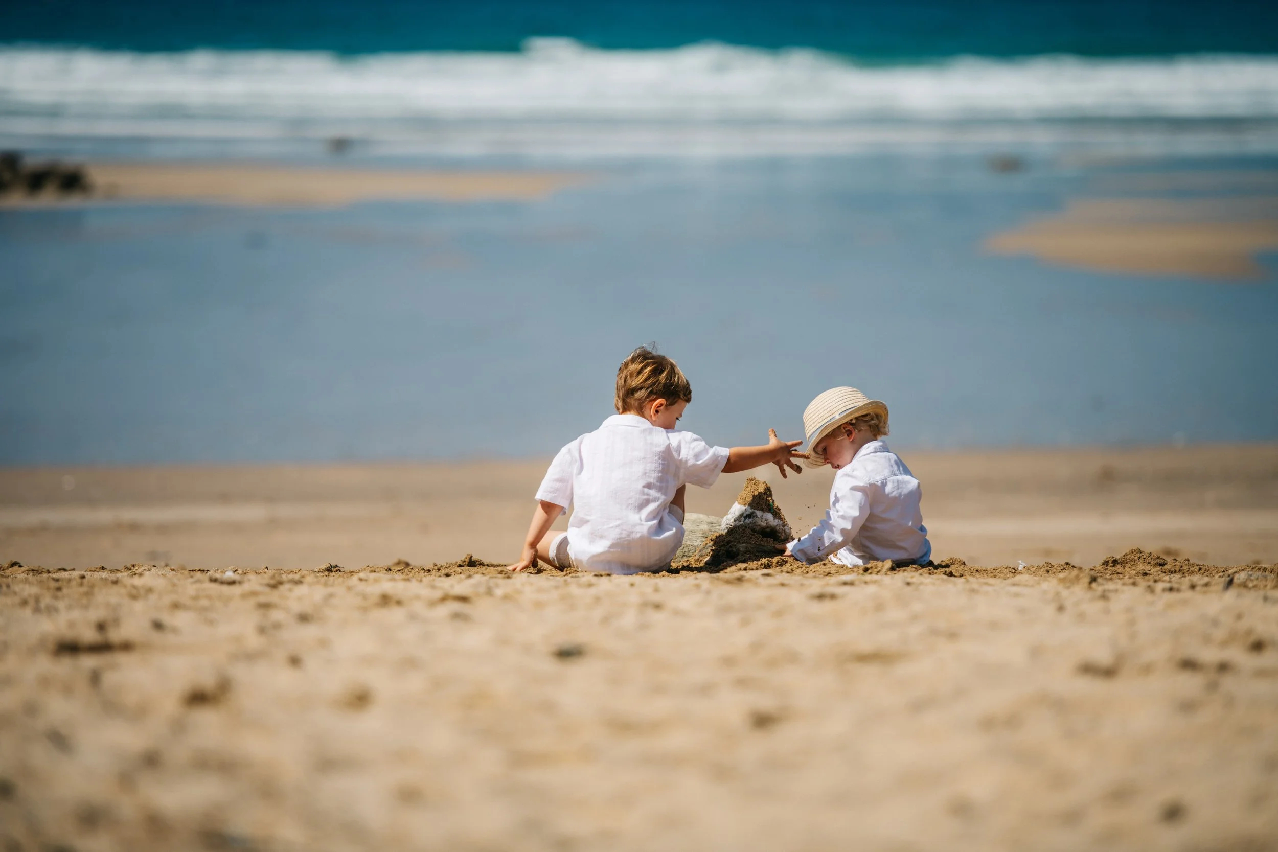 Two young children, wearing white clothes, playing and building a sandcastle on the beach near the ocean, with a sandy shore and waves in the background.