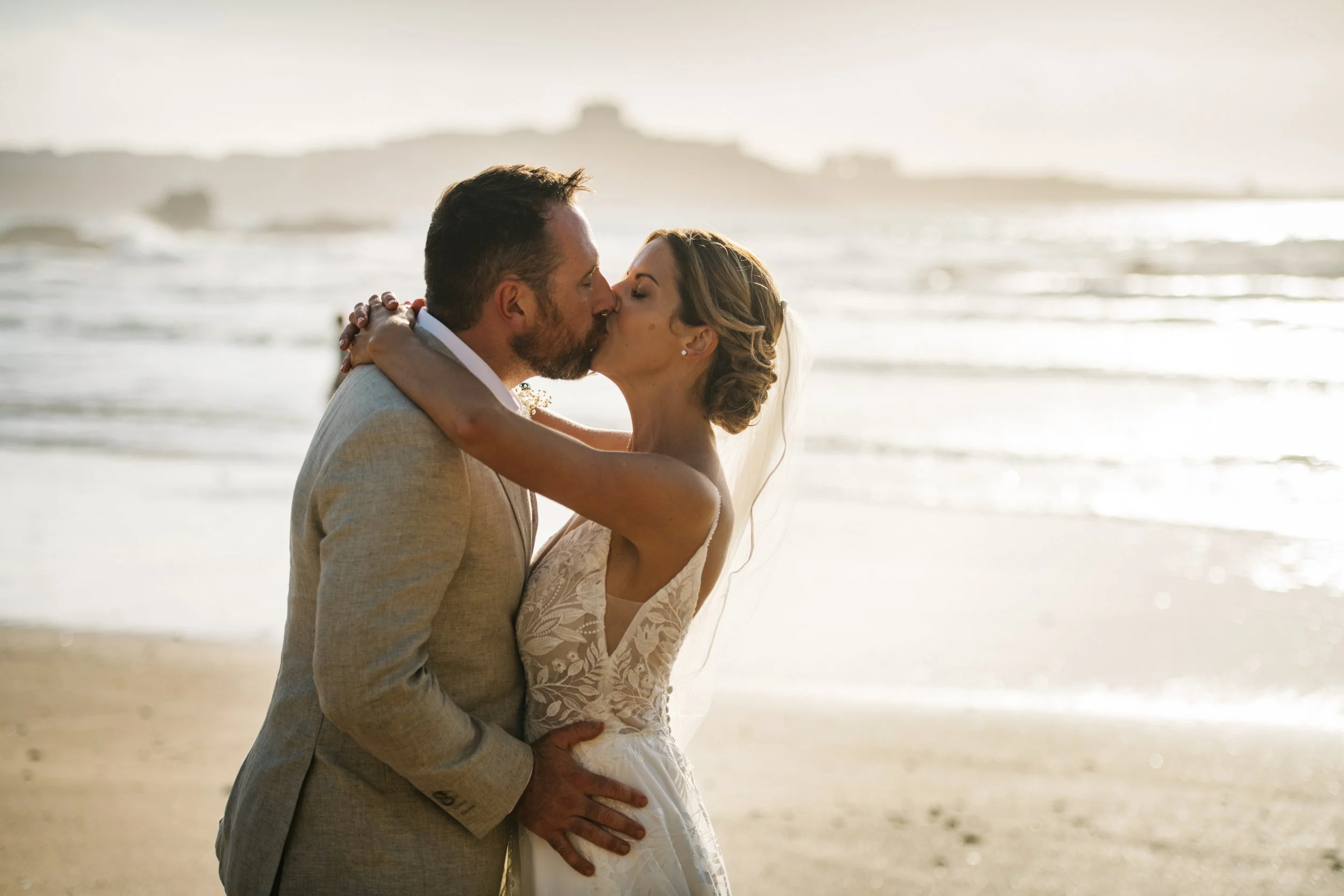 A bride and groom kissing on the beach at sunset, with waves in the background.